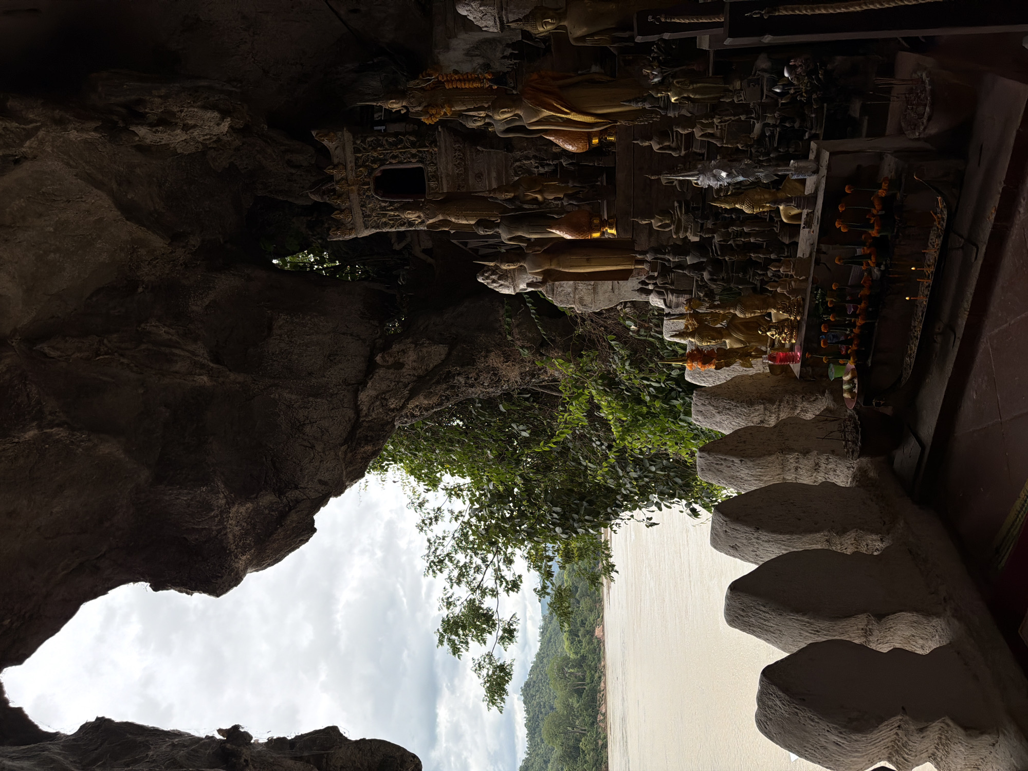This image depicts a scene within a cave in Muangkeo, Laos. The cave entrance frames a view of a wide, calm river bordered by lush, green hills under a cloudy sky. Inside the cave, a collection of Buddha statues of various sizes and materials are arranged on tiered platforms, some adorned with floral offerings and incense sticks. A tiered, ornate shrine structure is visible behind some of the statues. The cave interior is dimly lit, with the light primarily coming from the entrance and some small lamps illuminating the statues. The foreground features a textured, low wall with decorative elements, and a small portion of a red mat is visible on the floor. The overall atmosphere is serene and spiritual.