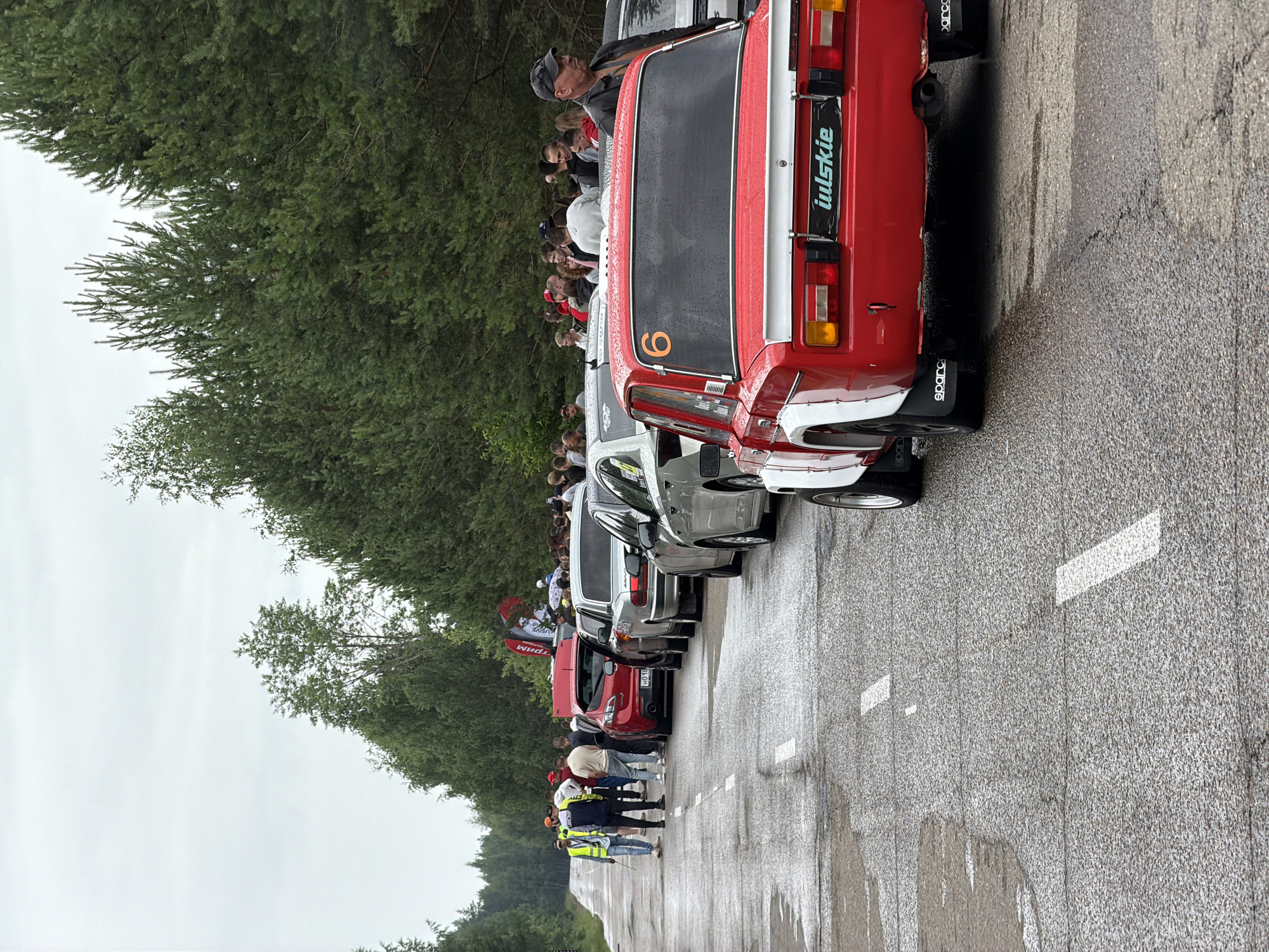 The image shows a line of cars on a wet asphalt road bordered by dense pine trees. The sky is overcast. In the foreground, a red vintage car with white trim is prominently featured. It has the number "6" in orange on its rear window and a sticker that reads "iulskie" on the rear bumper. The car has racing modifications, including wide fender flares and a rear spoiler. Several people are standing on the side of the road, some watching the cars, others in conversation. Further down the road, more cars are parked in a line, and a group of people are gathered. The scene appears to be a car event or competition. The presence of numerous pine trees suggests a location outside of a dense urban area, likely in a rural or suburban setting. Given the specific details within the image, such as the car models and overall atmosphere, it is possible this scene depicts an event in or near the city of Chaikovsky, Russia, which is known for its proximity to natural landscapes and forests.