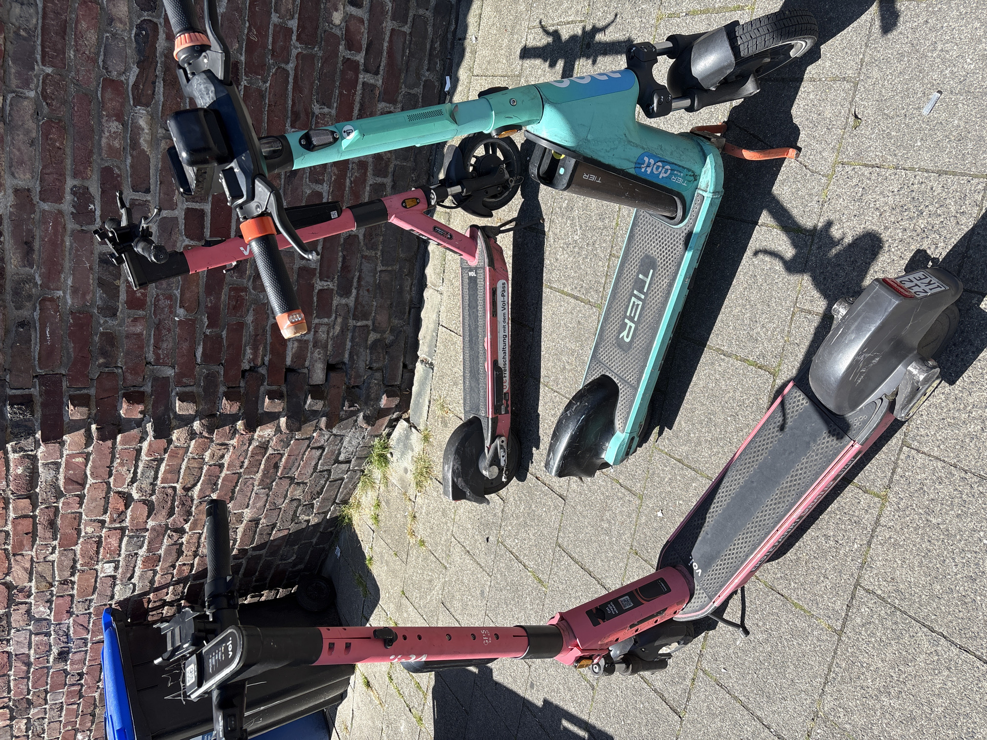 Three electric scooters are parked on a paved sidewalk in front of a brick wall. The scooter on the left is pink and has "voi." written on the footboard. The middle scooter is also pink and has a "Voi" logo on the stem. The scooter on the right is teal and has "TIER" and "dott" written on it. A blue-lidded trash bin is partially visible to the left of the pink scooter. The scene is illuminated by sunlight, casting shadows. The presence of Voi and Tier/Dott scooters suggests a city that has these shared mobility services, and the brickwork and paving are consistent with an urban environment. Based on the scooters and the brick architecture, this image was likely taken in Mönchengladbach, Germany, which features both Voi and Tier scooter services.