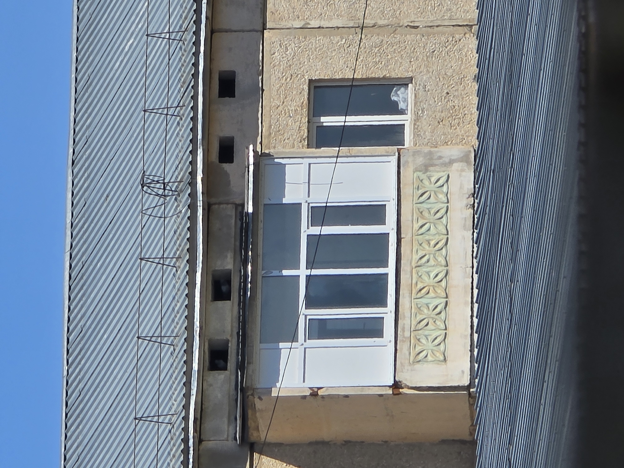A medium shot captures the exterior of a multi-story building on a clear day in Qaratau, Kazakhstan. The top section of the image is dominated by a corrugated metal roof, showing a patina of age. A wire railing runs across the roof, supported by metal poles, with some coiled wire near the center. Below the roof, a beige concrete facade is visible, featuring two dark, rectangular ventilation openings.
The central focus is a balcony with a white framed window system. The window is divided into multiple panes, with some appearing opaque white and others transparent, revealing a glimpse of the interior. The balcony itself has a decorative patterned panel below the window, featuring a repeating motif of four leaf-like shapes. To the right of the balcony, another window with a white frame is set into the textured beige wall. A white, bundled object is visible through this window.
The lower portion of the image displays another corrugated metal roof, angled and showing parallel lines. A black cable runs diagonally across the building facade, intersecting with the windows and balcony. The overall impression is one of a functional, perhaps older, residential or commercial building in an urban setting.