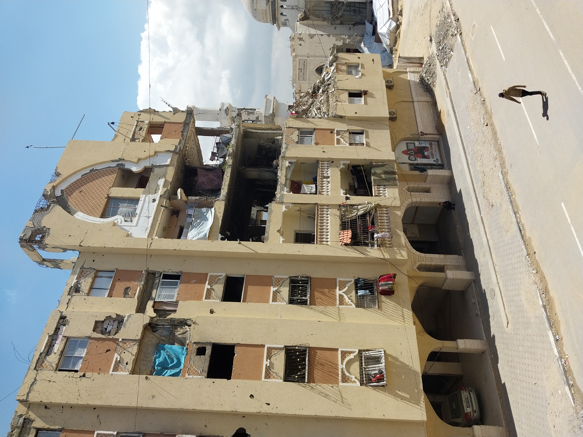 This image depicts a street scene in Khan Yunis, Palestinian Territory, dominated by a heavily damaged multi-story building under a partly cloudy sky. The main structure, beige with terracotta accents, exhibits extensive destruction, including large missing wall sections, exposed rebar, shattered windows, and numerous pockmarks across its facade. Clothes are visible hanging from some windows and balconies. On the ground floor, arched open spaces reveal a silver car parked on the left, and a "YASEEN GAMES" sign with a Squid Game motif on the right. A paved street with white lane markings runs horizontally across the foreground, where a solitary individual in a light brown jacket and dark pants walks from right to left. Another individual is seated on the sidewalk near the building's base. In the background, beyond the damaged building, a white mosque with a dome and minaret, also damaged, is visible amidst further rubble and several white tent structures on sandy ground.