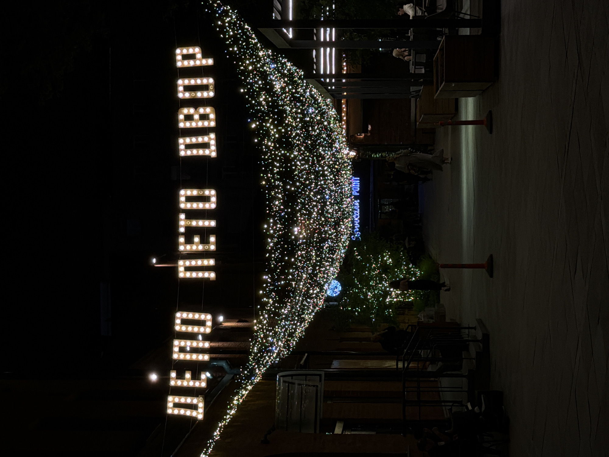 The image depicts a nighttime scene at a public space in Moscow, Russia. A lighted sign reading "Депо. Лето. Двор" (Depot. Summer. Courtyard) is prominently displayed. A canopy of small lights spans the space. Several people are present, seated at tables and walking. The area has paved surfaces and architectural elements.