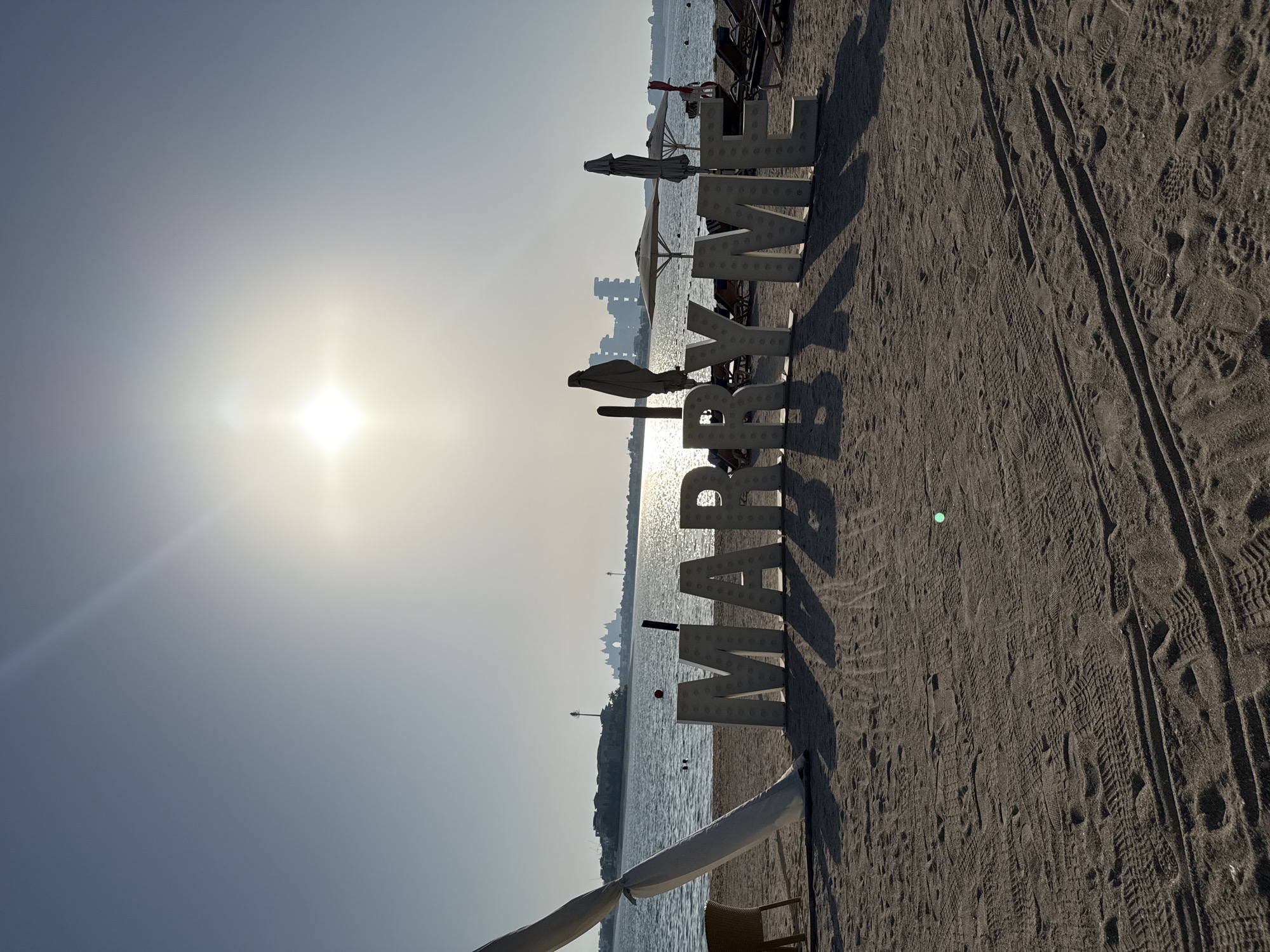 The image captures a beach scene in Dubai, United Arab Emirates, with large white, illuminated block letters spelling out "MARRY ME" positioned on the sand. The letters are casting long shadows on the beach, indicating it is either early morning or late afternoon due to the sun's position. The sun itself is a bright, almost blinding orb in the sky, slightly off-center to the right, creating a lens flare effect. In the background, the calm, blue water of the Persian Gulf is visible, with distant buildings and structures on the horizon, characteristic of Dubai's skyline, possibly The Palm Jumeirah. Several beach chairs and umbrellas are scattered on the sand behind the "MARRY ME" sign. A white tent-like structure with a draped fabric is partially visible on the left side of the frame. The sand is textured with footprints and tire tracks. There are no people clearly visible in the foreground or interacting with the sign, but there are faint figures in the water.