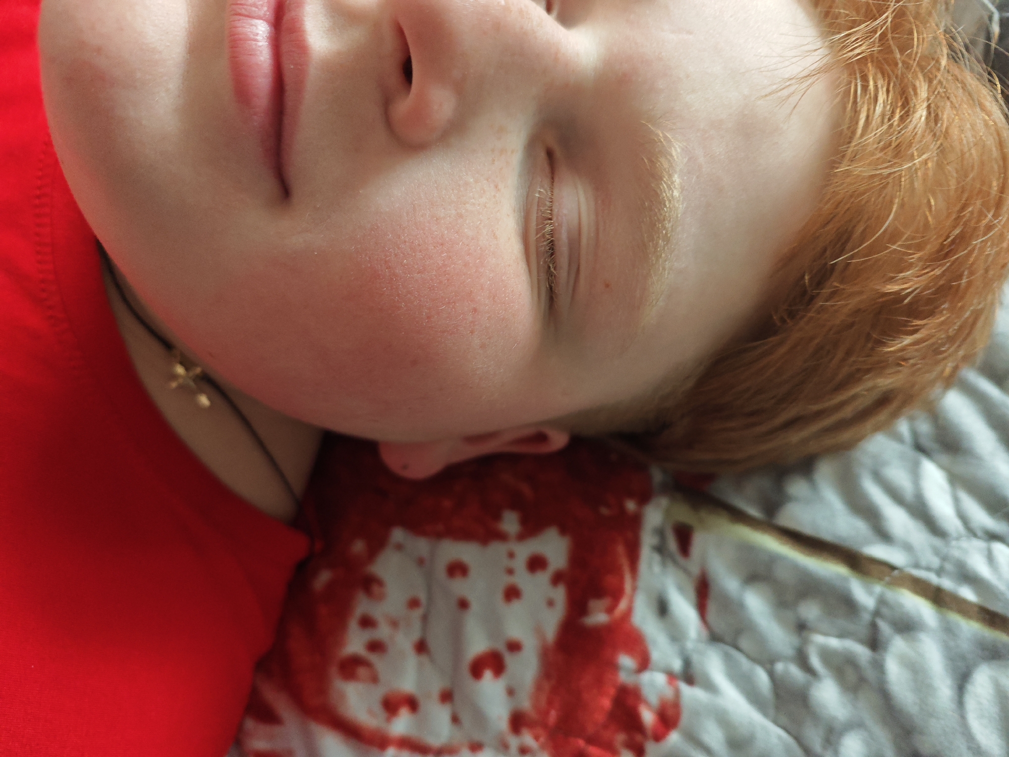 The image shows a close-up of a person with fair skin, red hair, and closed eyes. The person is lying on a patterned gray and white surface, possibly a quilt. There is a reddish-brown substance resembling blood near the person's head and ear. The person wears a red garment and a necklace with a cross. The location, Markovskii, Russia, is provided as context but is not directly discernible from the image.