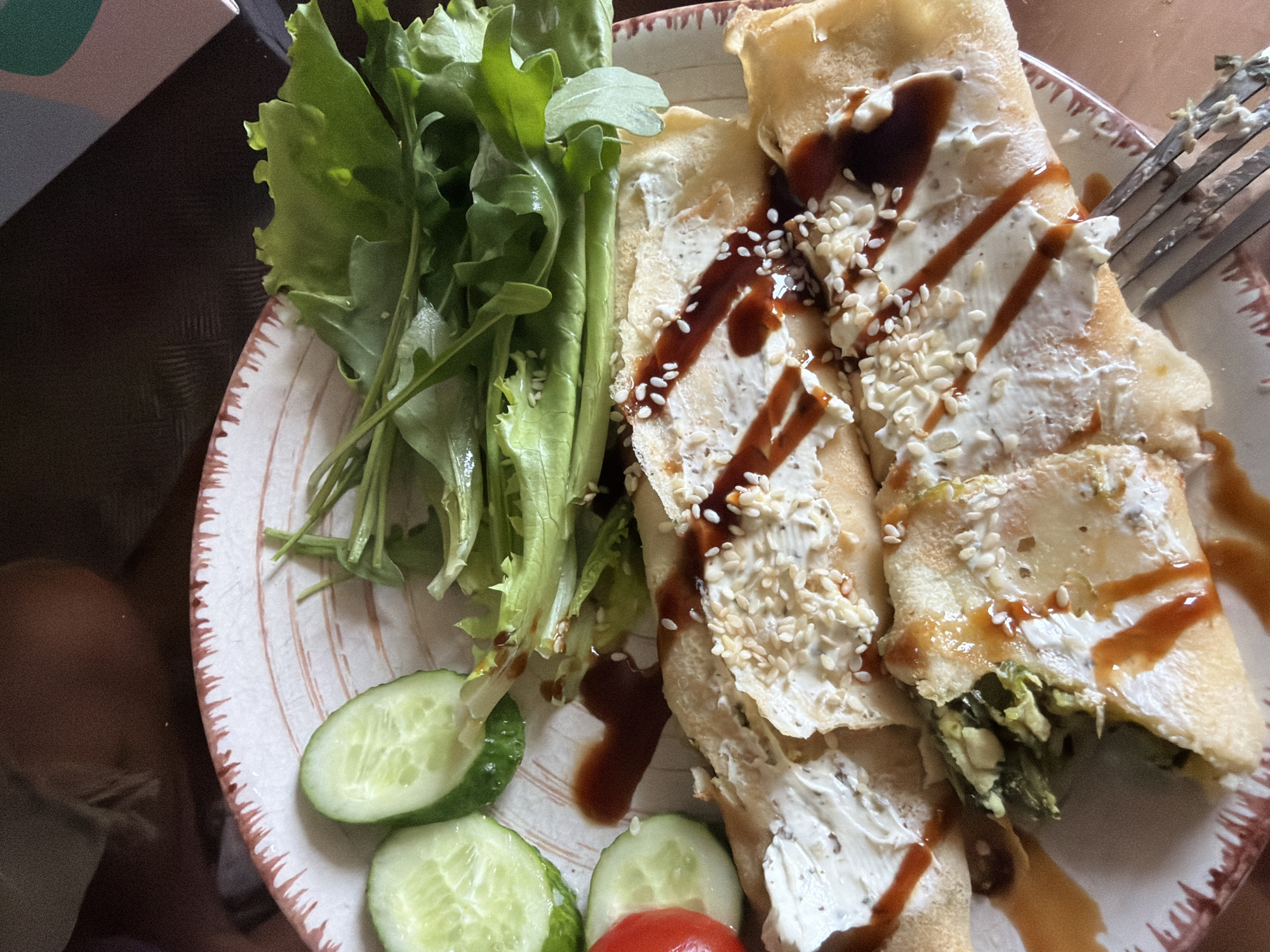 The image shows a plate of food on a dark brown surface. The plate is white with a red-brown decorative rim and contains crepes filled with a white creamy substance and possibly some green ingredients. The crepes are drizzled with a dark sauce and sprinkled with sesame seeds. To the left of the crepes are sliced cucumbers and a halved cherry tomato. To the top of the crepes, there is a pile of green leafy vegetables, possibly lettuce and arugula. A fork is visible on the right side of the plate, with some food on its tines. The background is dark and out of focus. There is no visible indication of the location being in Kyiv, Ukraine.