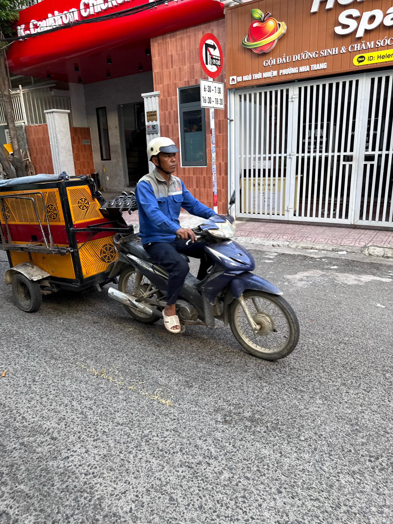 In Nha Trang, Vietnam, a man wearing a light-colored helmet and a blue and gray uniform rides a dark blue motorcycle. He is pulling a yellow and red trailer with a black cover. The trailer has a cage-like structure and appears to be used for transport. The man is wearing white sandals and dark pants. He is on a paved street with some debris and a painted yellow line. In the background, there are buildings with signs. One sign displays a red apple logo and text in Vietnamese, indicating "SPA" and a street address. Another sign has a red circle with a diagonal line and an arrow, suggesting a traffic regulation. The scene appears to be during daylight hours.