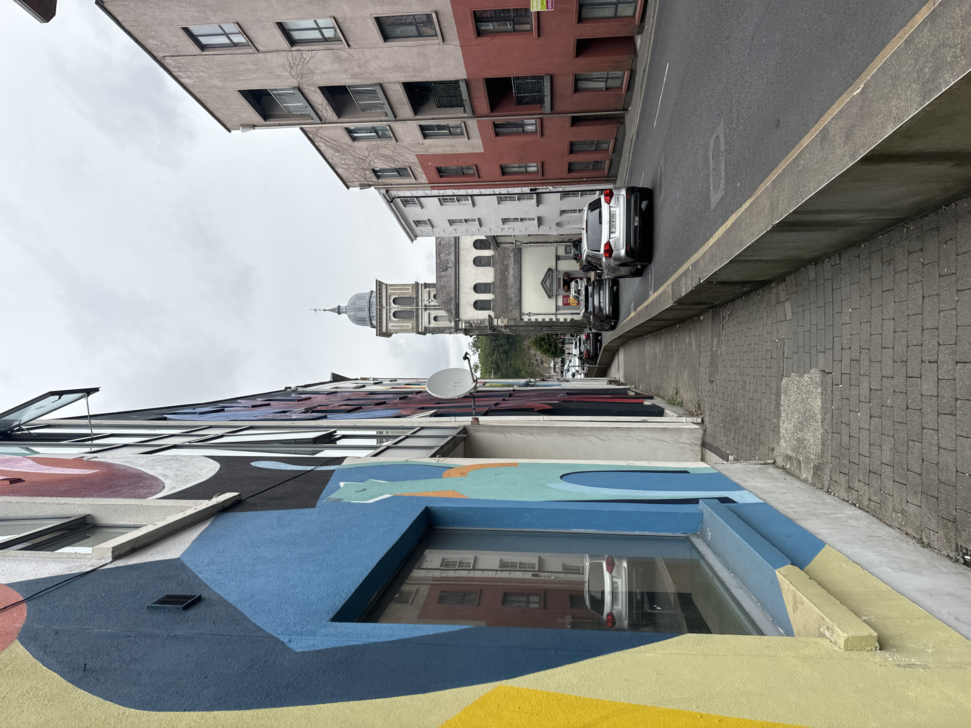 The image depicts a street scene in Waterford, Ireland, specifically on The Mall, with a view towards the tower of Christ Church Cathedral. On the left side of the image is a building with vibrant, abstract mural art. The mural features large, colorful shapes in yellow, blue, red, and black, with a stylized teal cat-like figure visible. A large window reflects a silver car parked on the opposite side of the street.
The street itself is paved with grey brickwork for the pedestrian walkway and asphalt for the road. A concrete barrier with a yellow stripe runs along the edge of the walkway, separating it from the road. Several cars are driving on the road, including a silver SUV in the foreground with license plate 241-W-494.
Across the street, a row of buildings of varying heights and colors line the road. The buildings are predominantly in shades of pink, red, and white, with windows and balconies. In the distance, the prominent tower of Christ Church Cathedral rises above the other structures, its dome silhouetted against a cloudy sky. The overall atmosphere is overcast, suggesting a typical Irish day. No people are clearly visible or engaged in any specific actions within the frame.