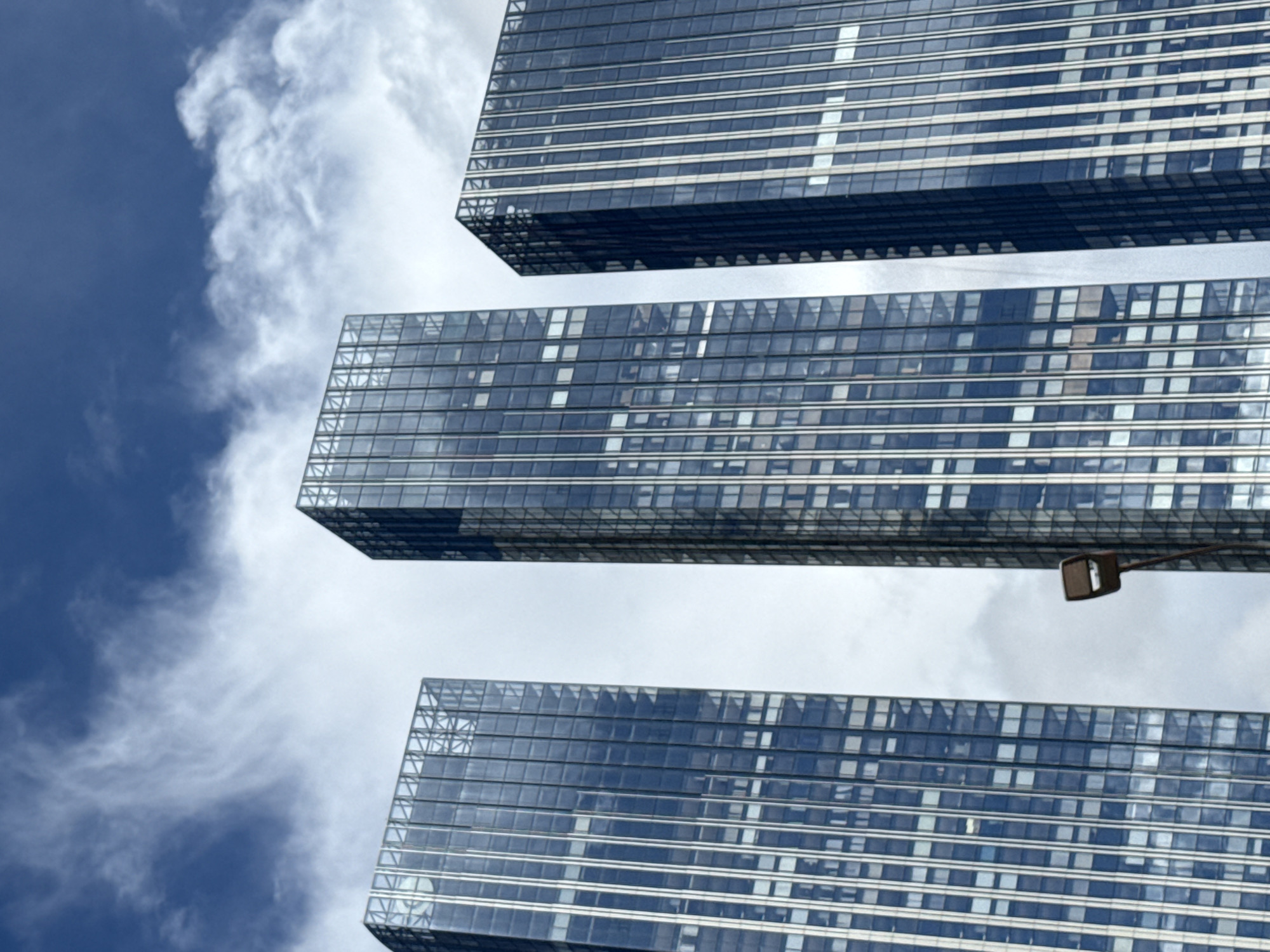 The image shows a low-angle view of three modern glass skyscrapers against a cloudy blue sky. The buildings are tall and rectangular, with repeating patterns of windows and vertical structural elements. In the foreground, a single lamppost with a rectangular fixture extends upwards from the bottom left of the frame. The scene appears to be an urban environment, likely a business district. The architecture suggests a contemporary setting. This image was captured on May 21, 2023. The specific location is the Moscow International Business Center, also known as Moscow City, a developing business district in western Central Moscow, Russia.