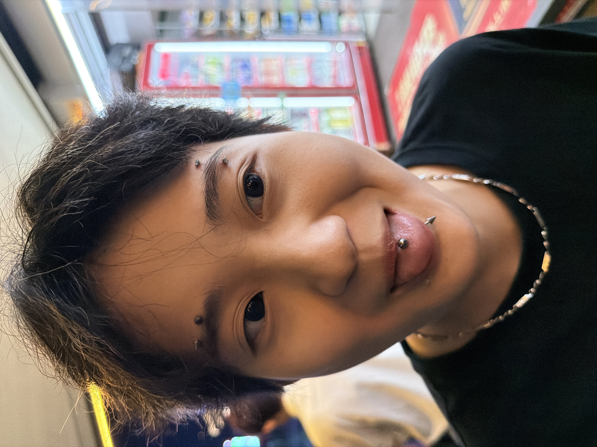 The image features a young East Asian individual in a close-up, front-facing shot, smiling broadly and extending their tongue. They have short, dark, somewhat disheveled hair. Their left eyebrow (viewer's left) has two surface piercings: one with a spherical stud and one with a flatter, disc-like stud. Their right eyebrow (viewer's right) also has two surface piercings, both with spherical studs. A barbell tongue piercing, featuring a spherical end and a pointed conical end, is visible on their extended tongue. The individual is wearing a plain black t-shirt and a silver-toned chain necklace with alternating small beads and rectangular links.
In the blurred background to the immediate left of the subject, a second individual wearing a light-colored shirt is partially visible. The background to the right reveals an indoor commercial setting, specifically an aisle within what appears to be a convenience store or supermarket. Multiple tall, glass-fronted refrigeration units, illuminated by vertical fluorescent lights, contain various beverages and packaged goods. A red floor mat is visible on the ground in the foreground of the background. The scene is under artificial interior lighting. The location is in the city of Jiangbeicheng Subdistrict, China.