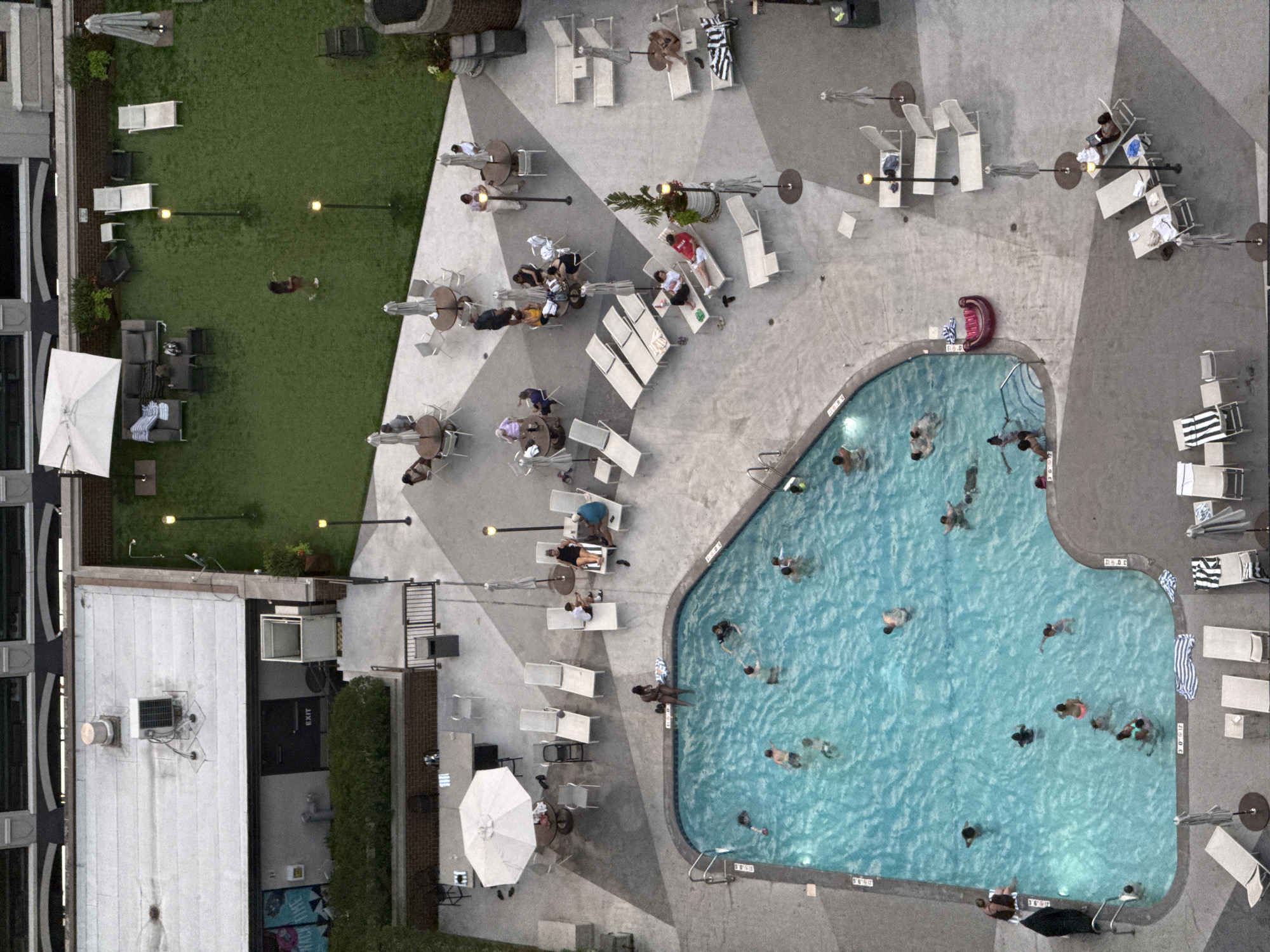 An aerial view captures a rooftop pool and lounge area at a building in Chicago, USA. The central focus is a large, kidney-shaped swimming pool filled with people swimming and interacting. Surrounding the pool are paved areas with lounge chairs, umbrellas, and tables, some occupied by individuals relaxing or conversing. Adjacent to the pool area is a grassy space with additional seating and umbrellas. A building with a rooftop patio and an outdoor seating area is visible. The overall scene depicts a vibrant urban leisure space on a bright day.