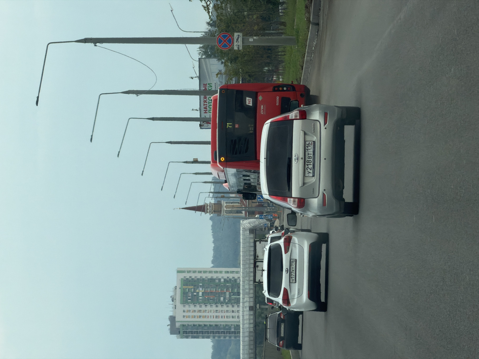 The image depicts a street scene in Kazan, Russia, with a focus on vehicles in traffic. In the foreground, a silver car is driving on a multi-lane asphalt road. Behind it, a white SUV with a Russian license plate (H177KT 716) is visible. Further ahead, a red bus with the number 71 on its front is seen, along with another vehicle. In the background, a tall modern building with colorful accents stands out, accompanied by a more traditional tower with a spire. Streetlights line the road, and a pedestrian overpass is also present. The sky is overcast, indicating a cloudy day. The scene suggests a typical urban commute or traffic situation.