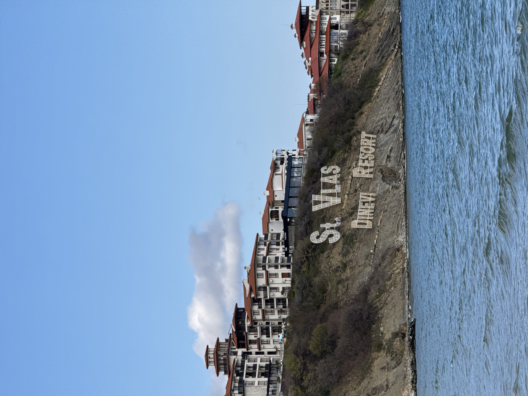 The image shows a view of Sveti Vlas, Bulgaria, from the water. Several multi-story buildings with red-tiled roofs cascade down a rocky hillside towards the water's edge. The buildings are white with dark window frames. Along the hillside, large white letters spell out "St. Vlas Dinevi Resort." The sky above is a clear blue with a few scattered clouds. The water is choppy with whitecaps, filling the right-hand side of the image.