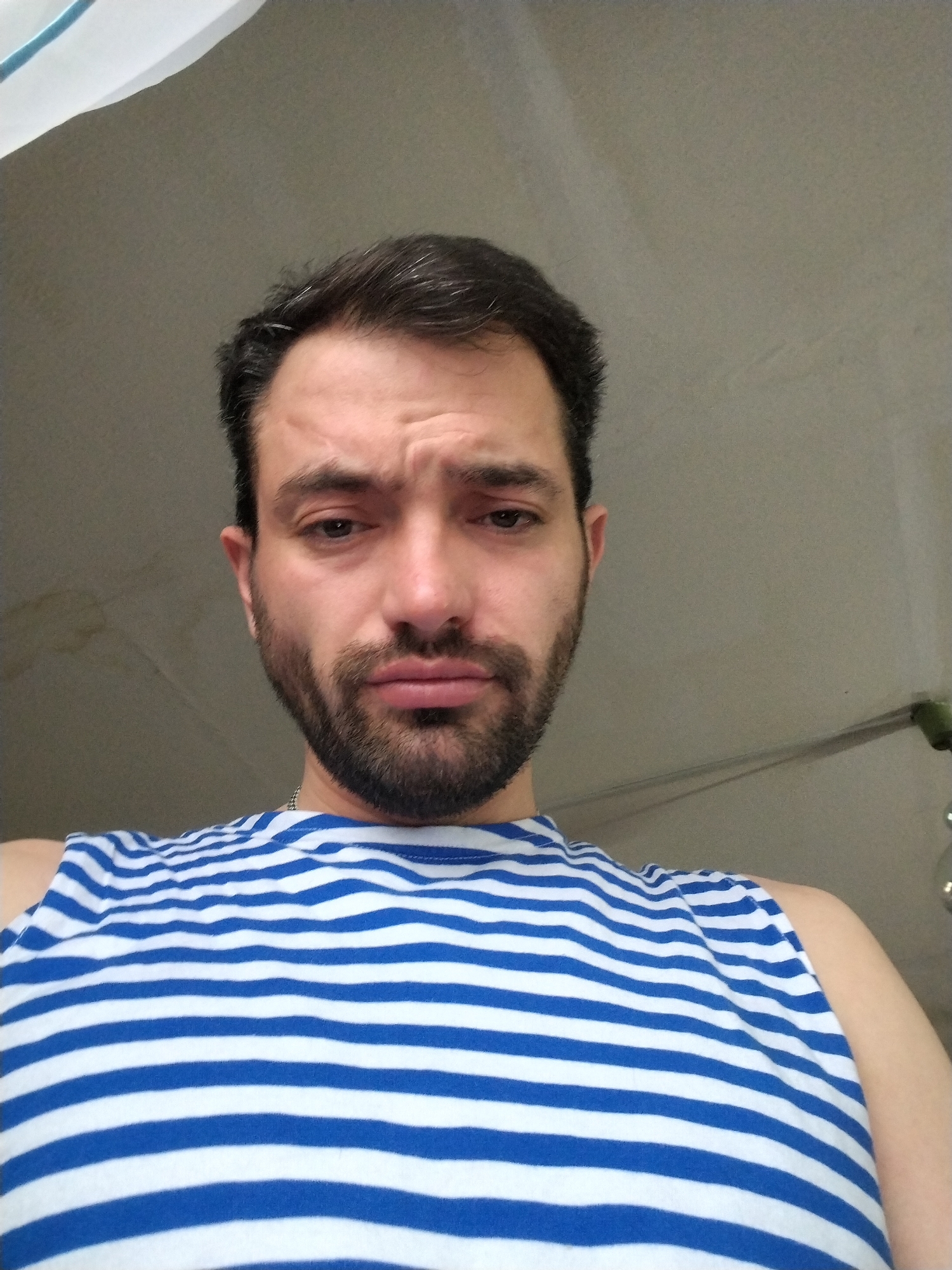 A man with dark hair and a beard is wearing a blue and white striped tank top. He has a concerned or sad expression on his face. The background is a plain, light-colored wall with some minor blemishes.
The location of the image is not provided.