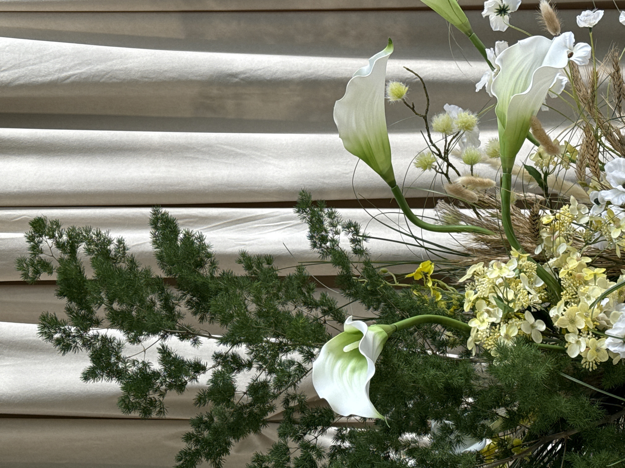 The image displays a detailed view of a floral arrangement positioned in an indoor setting. Prominently featured are three white calla lily-like flowers, each with a green stem and a spathe that transitions from green at the base to pure white at the wavy edges. One lily is centered on the left, another arches from the upper right, and a third curves upwards from the lower right. The arrangement includes a substantial volume of dark green, feathery foliage, resembling asparagus fern, which forms a dense background for the lilies. Clusters of small, light yellow or cream-colored flowers are visible throughout the lower and central parts of the arrangement, alongside thin green grasses and dried brown stalks, possibly wheat or other decorative grasses. Small, fuzzy, light green spherical plants on thin branches are also integrated. The background consists of light beige or off-white curtains, vertically pleated, creating shadows and depth. No individuals are present in the scene. The arrangement appears static, functioning as a decorative display. There is no visual information within the image to indicate its specific geographical location, including the city of Perm, Russia.