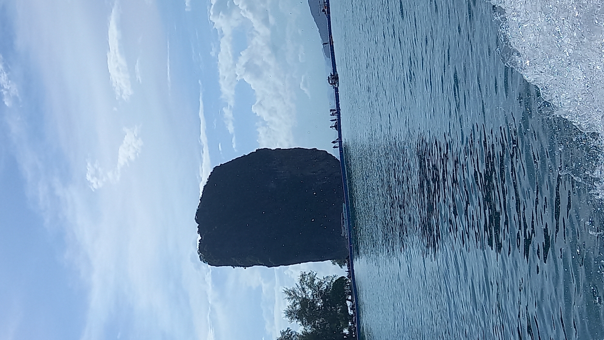 The image captures a scenic view in Ban Ao Nang, Thailand, featuring a prominent dark, vegetated limestone karst island near the shoreline. The foreground is dominated by turquoise water with subtle wave patterns. The sky is partly cloudy. A portion of a pier or structure lines the horizon, and several indistinct figures appear to be on it. Trees are visible on the shoreline, near the base of the karst island. The angle of the shot suggests it was taken from a boat or moving platform at water level.