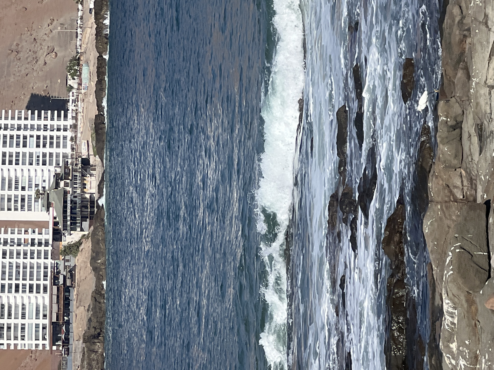 The image shows a coastal scene in Arica, Chile. On the left are several high-rise white buildings situated along a sandy beach and urban coastline. The right side of the image is dominated by a rocky shoreline battered by waves. The waves are crashing against the rocks, creating white foam and spray. The water transitions from deep blue in the center to a lighter, more turbulent hue near the shore.
