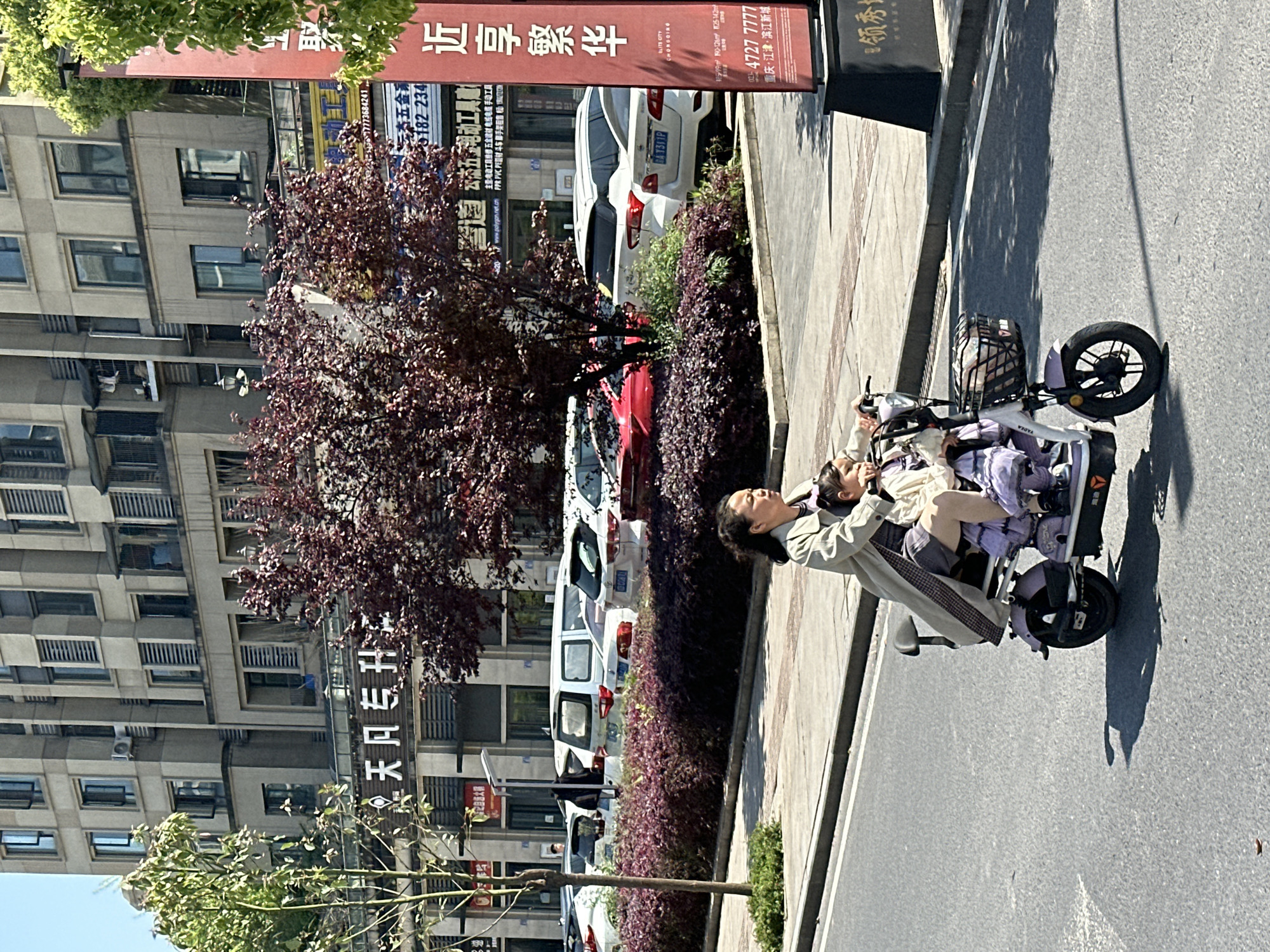 A woman and a child are riding an electric tricycle on a street in the city of Jiangjin, China. The woman is wearing a tan coat and the child is wearing purple and white clothing. The tricycle has a basket on the front. In the background, there are buildings, parked cars, and a sign with Chinese characters that translates to "Leading Show". The sign also has the phone number 4727 7777. A closer look reveals the location as Chongqing Jiangjin Binjiang New City.
