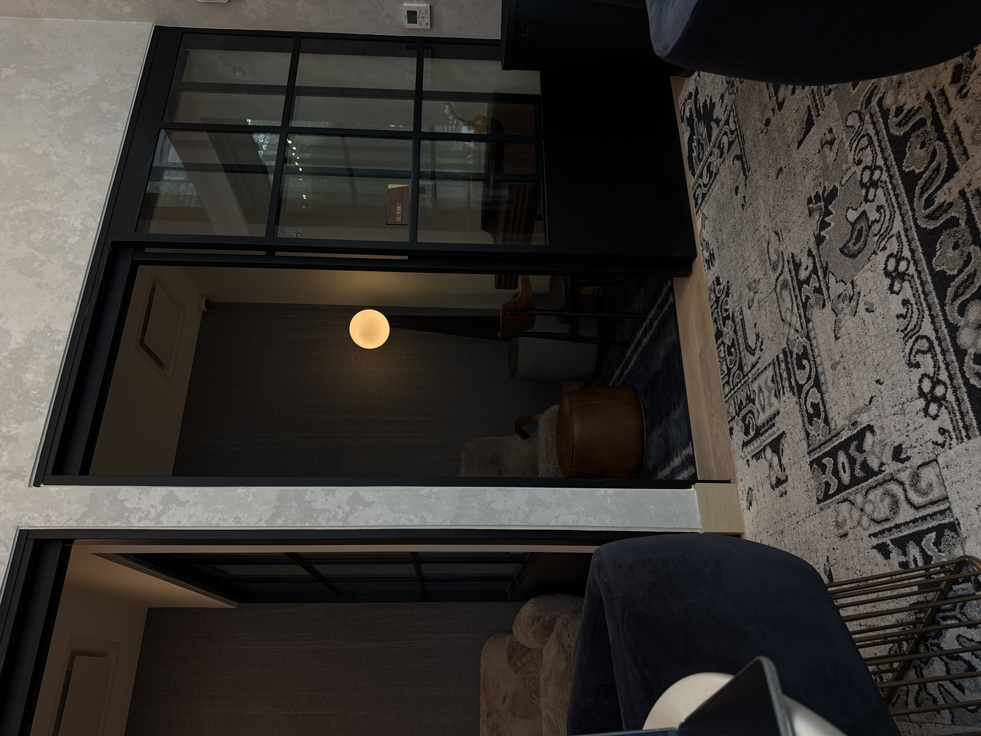 An interior scene depicts a modern room, possibly a lounge or waiting area, separated by a striking black-framed glass partition from another room beyond.
In the foreground, to the right of the partition, a light-colored, textured wall, resembling stucco or concrete, runs vertically. To the left, a portion of a dark blue upholstered chair is visible, along with a light-toned rug featuring a prominent dark, intricate pattern, suggestive of a Persian or geometric design. A metal rack with horizontal bars is partially visible beneath the chair, and a small, light-colored object rests on the chair.
Through the clear panes of the glass partition, a second, dimly lit room is visible. The dominant feature in this room is a glowing spherical light fixture, casting a warm yellow light against the dark, textured walls. Within this room, a brown cylindrical object, possibly a pouf or a piece of luggage, sits on a fluffy, light-colored rug or cushion. A beige upholstered chair and a wooden chair with a slatted back are also discernible. On one wall, a white control panel, likely for HVAC, is mounted. In the upper section of the room, seen through the glass, a shelf holds a vase containing white decorative elements.
No people are present in the scene, which appears to be static. The atmosphere is calm and inviting, with the prominent lamp suggesting evening or low-light conditions. The visible text "ROOM 2" is seen on a brown sign or block within the distant room, indicating its designation. The overall aesthetic is contemporary and sophisticated, aligning with a modern office, hotel, or upscale residential common area in Austin, United States of America.