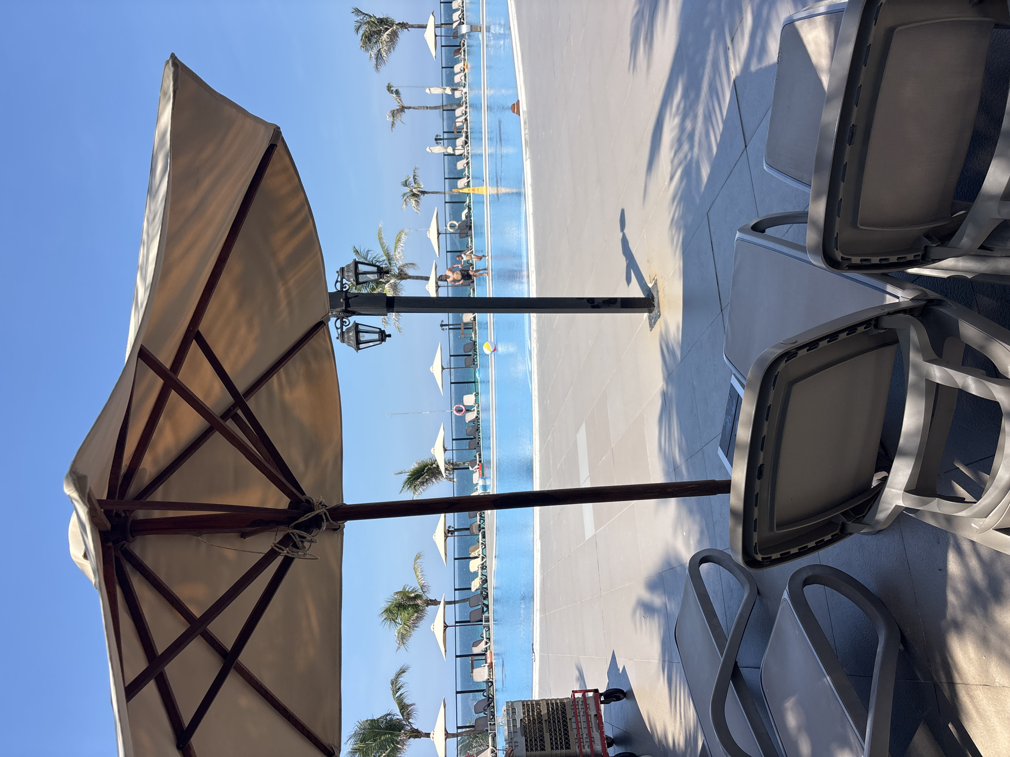 An outdoor swimming pool area is depicted under a clear blue sky. In the foreground, several empty lounge chairs are arranged on a paved surface, with shadows from palm fronds cast across them. A large, beige patio umbrella with a wooden frame dominates the upper portion of the image. Behind the lounge chairs, a bright blue swimming pool stretches across the middle ground. Along the far side of the pool, a row of lounge chairs and umbrellas are visible, with palm trees and the ocean in the background. Two people, a man and a child, are standing in the pool, with the child appearing to be running. A yellow cone and a life preserver are also in the water. To the left, a cart with stacked crates is partially visible. A black lamppost with two lantern-style lights stands near the center of the frame.