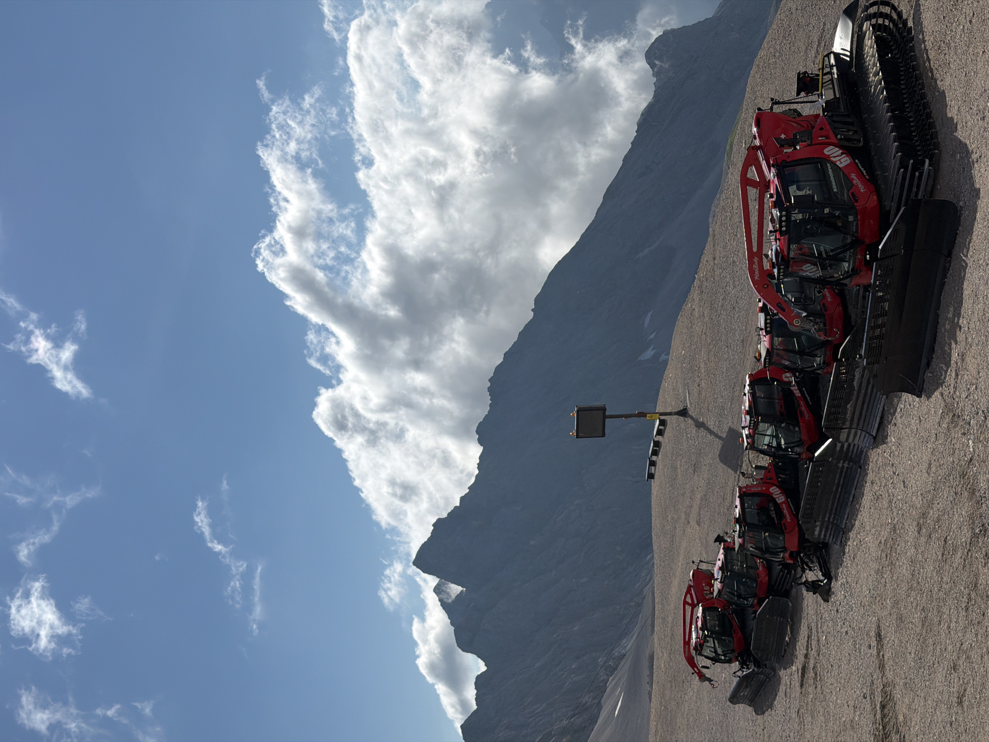 This is a medium-shot, vertical photograph taken from a slightly elevated position, looking down at a line of red snowcats parked on a gravelly surface on a mountainside. The snowcats are the primary subjects in the foreground, with their tracks and front plows visible. They are positioned diagonally from the bottom left corner, extending towards the center of the frame and then curving slightly to the right. Each snowcat has a cab with tinted windows and various lights on top. Some of the snowcats have branding visible, such as "Kässbohrer" and "6040."
In the middle ground, to the right of the snowcats, a signpost with a dark square display and a yellow warning sticker stands on a pole. Two benches are placed behind the signpost. Further back, the terrain slopes upwards, composed of gravel and some patches of snow.
The background is dominated by a vast mountain range with jagged peaks. Dark gray and rocky mountains fill the mid-to-far background, with some clouds obscuring parts of them. The sky above is a vibrant blue, with scattered white and gray clouds, some of which are large and billowy, partially obscuring the upper portions of the mountains. The overall lighting suggests it is daytime with bright sunshine.
The location is Garmisch-Partenkirchen, Germany, specifically at the Zugspitze. The scene depicts a mountainous, high-altitude environment, likely a ski resort or a service area for mountain operations, given the presence of the snowcats. There are no people visible in the image.