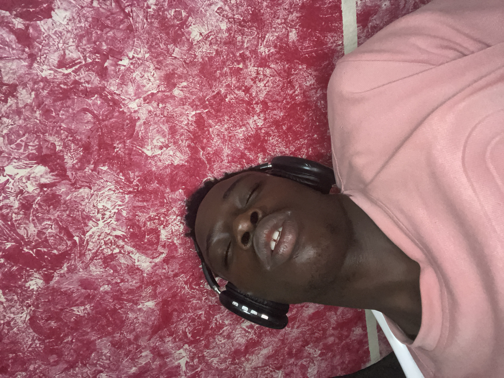 A person is shown wearing headphones and lying down. The background consists of a red textured wall. No information regarding location or city of Obosi, Nigeria, is discernible. The person wears a pink garment.