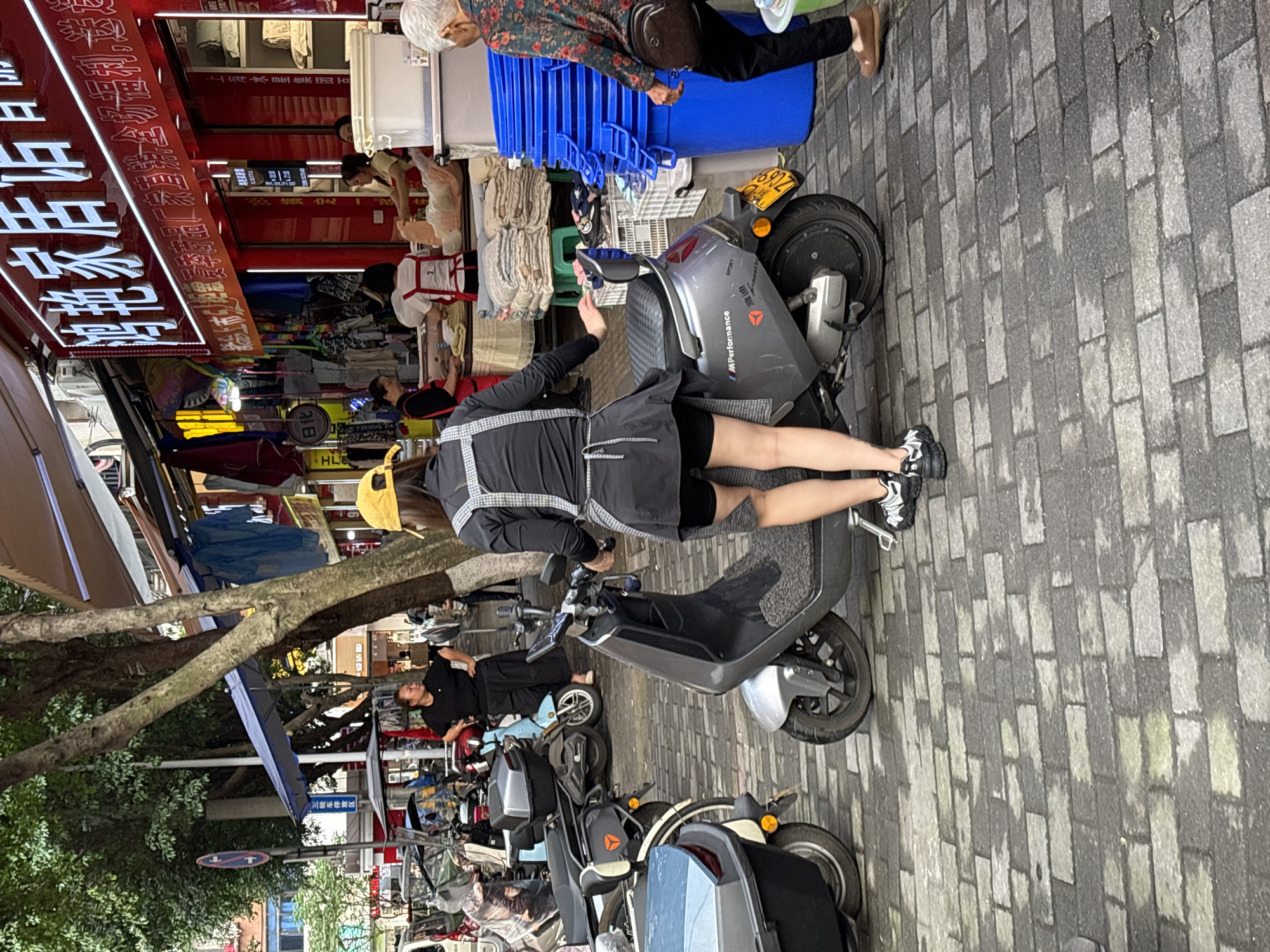 The image shows a street scene in Jiangjin, China. In the foreground, a person wearing a black dress and apron is standing beside a gray electric scooter. Behind the person, there are several other parked scooters. In the background, there are several people, including a person with gray hair wearing a floral shirt. There is a shop with a red sign with Chinese characters in the upper-left corner. The street is paved with gray bricks. There is a tree on the left side.