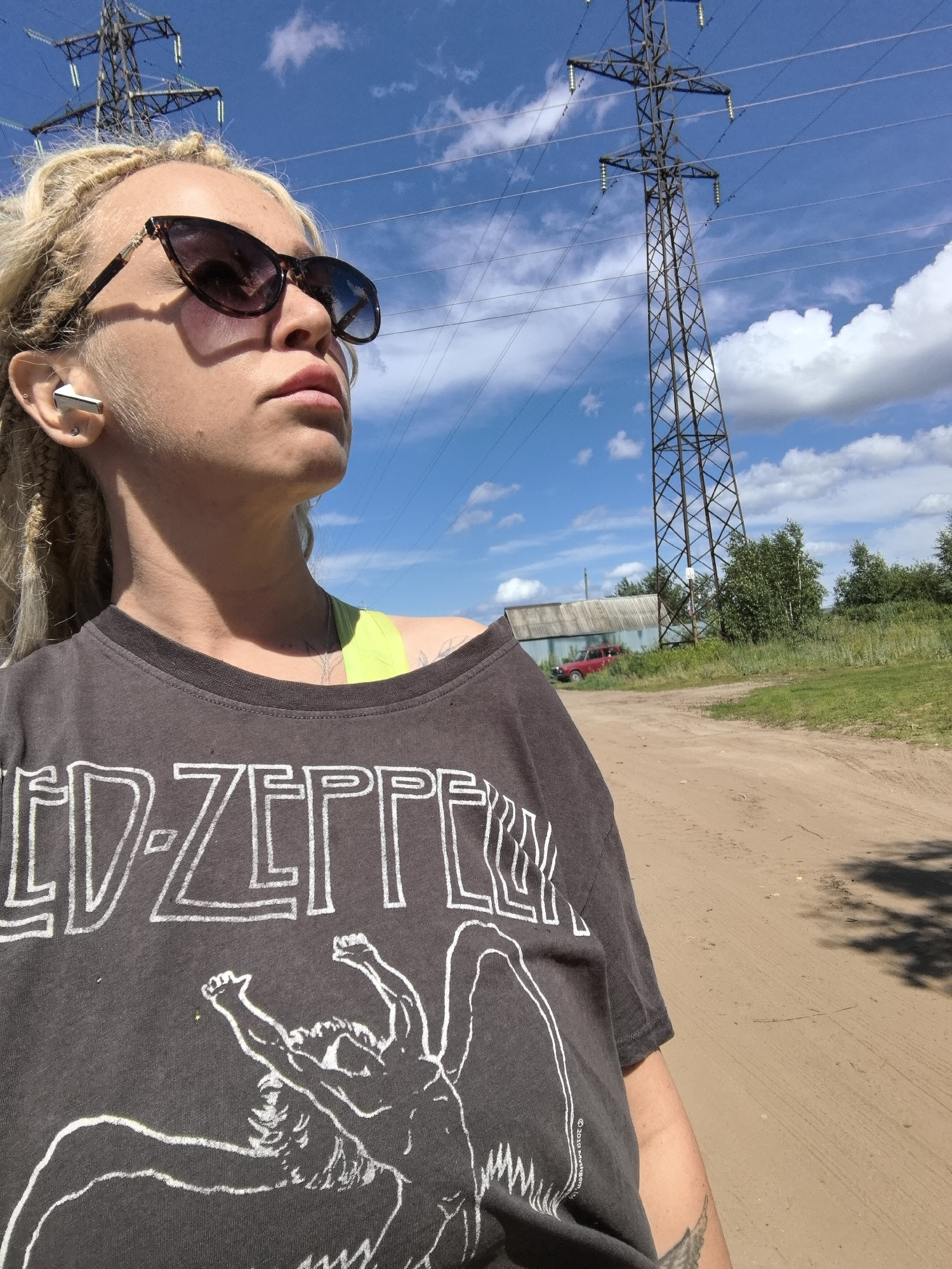 A woman with blonde dreadlocks and sunglasses takes a selfie outdoors on a sunny day. She is wearing a dark grey t-shirt with a white Led Zeppelin logo and graphic of a winged figure. She has on white earbuds and a silver necklace. In the background, a tall electrical tower dominates the sky, with wires stretching across. A dirt road stretches out in front of her, with green grass and trees lining the sides. In the distance, a weathered building and a red car are visible. The sky is a vibrant blue with fluffy white clouds. The overall mood is casual and relaxed, suggesting a daytime activity in a rural or semi-rural setting.
