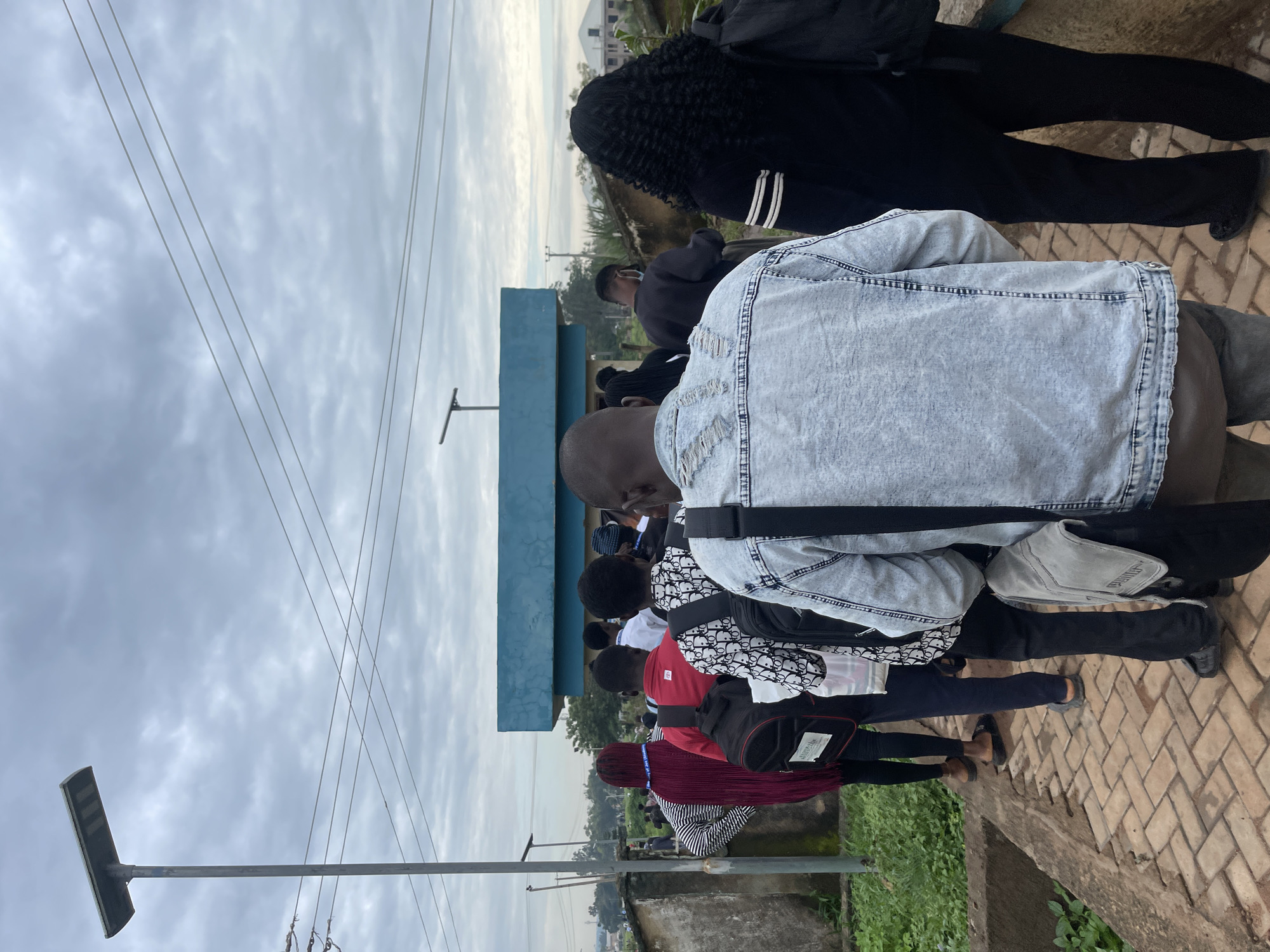 A group of young adults, predominantly of West African descent, are visible from the rear, standing in a queue on a paved walkway. They appear to be outside an institution, possibly a school or administrative building, given the presence of a blue-accented structure in the background and a solar-powered lamppost. The sky is overcast, suggesting a cloudy day. The individuals are dressed in casual attire, with several carrying backpacks. The environment seems to be within a semi-urban or rural setting in Rafin Par, Nigeria, indicated by the vegetation and distant buildings. The individuals are facing away from the camera, and their postures suggest they are waiting patiently. The walkway is constructed of interlocking brick pavers.