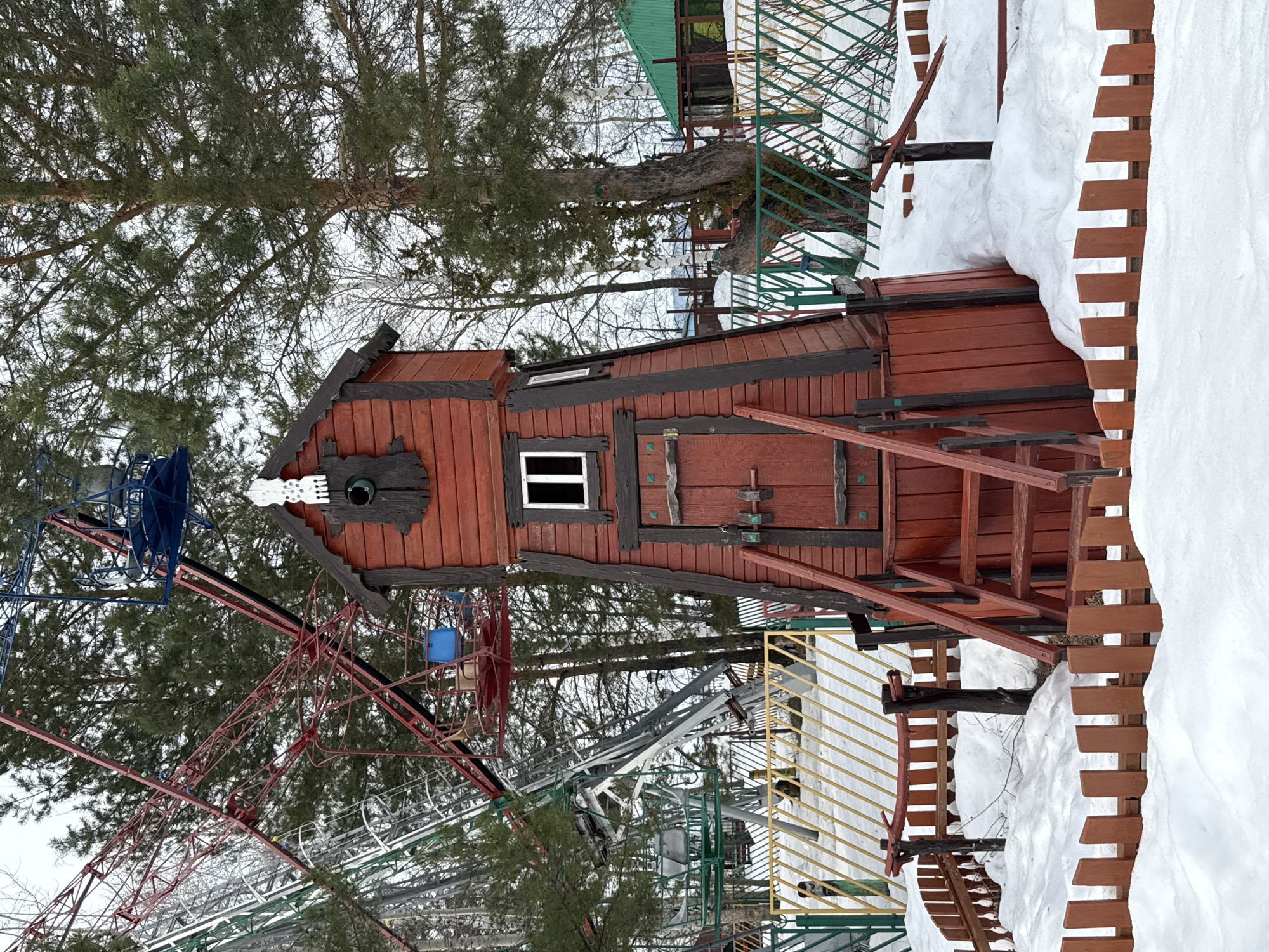 This image depicts a snow-covered outdoor scene in Chaikovsky, Russia, within what appears to be an amusement park or recreational area. Central to the frame is a multi-story, reddish-brown wooden structure, resembling a rustic tower or playhouse, featuring dark brown trim and a conical roof. It has a wooden door with visible metal hardware, a white-framed rectangular window above it, and another small white-framed window on its right side. The apex of the structure includes a round, dark-framed opening surmounted by an irregular white decorative element. Reddish-brown wooden stairs with handrails provide access to the door. To the left, a stationary Ferris wheel is visible, displaying red, green, and silver metal framework along with blue and grey gondolas, set against evergreen pine trees and bare deciduous trees. The ground in the foreground is covered in uneven snow, partially obscuring a low wooden picket fence of reddish-brown planks. A green and yellow metal fence with a diagonal pattern runs in the midground. The sky is overcast. No people are present.