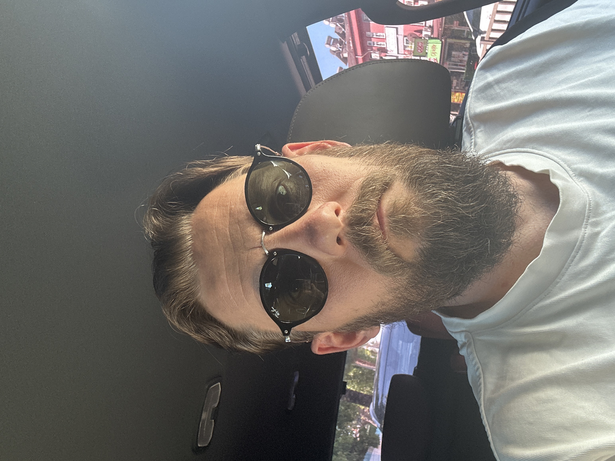 A man with a salt-and-pepper beard and sunglasses is in a car, likely in Fethiye, Turkey. The reflection in the car window shows a streetscape with buildings featuring signs with what appears to be Turkish script. The man is wearing a white shirt and the interior of the car is dark, with a headrest visible behind him. The sunglasses obscure the man's eyes.
