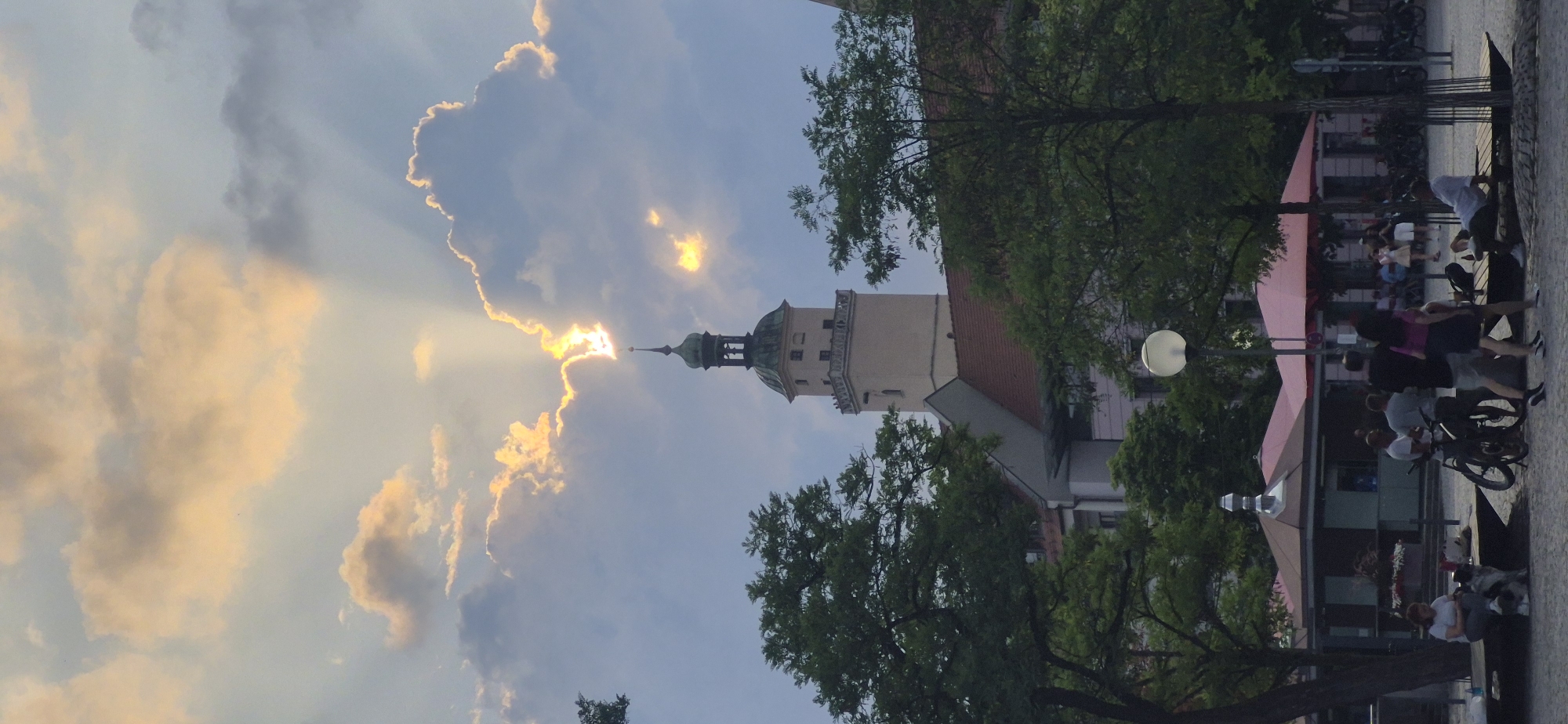 The image captures a view of the New Town Hall tower in Ingolstadt, Germany, during a cloudy sunset. The sun's rays break through the clouds, creating dramatic lighting effects behind and around the tower's spire. The tower itself is light-colored with a green-capped roof and multiple small windows. In the foreground, lush green trees partially obscure the view of the tower and surrounding buildings, which have red tile roofs. Below the trees, a paved area shows people sitting on benches and standing around, with some bicycles visible. There are also some small kiosks or market stalls with red awnings. The overall atmosphere suggests a public square or outdoor seating area.
