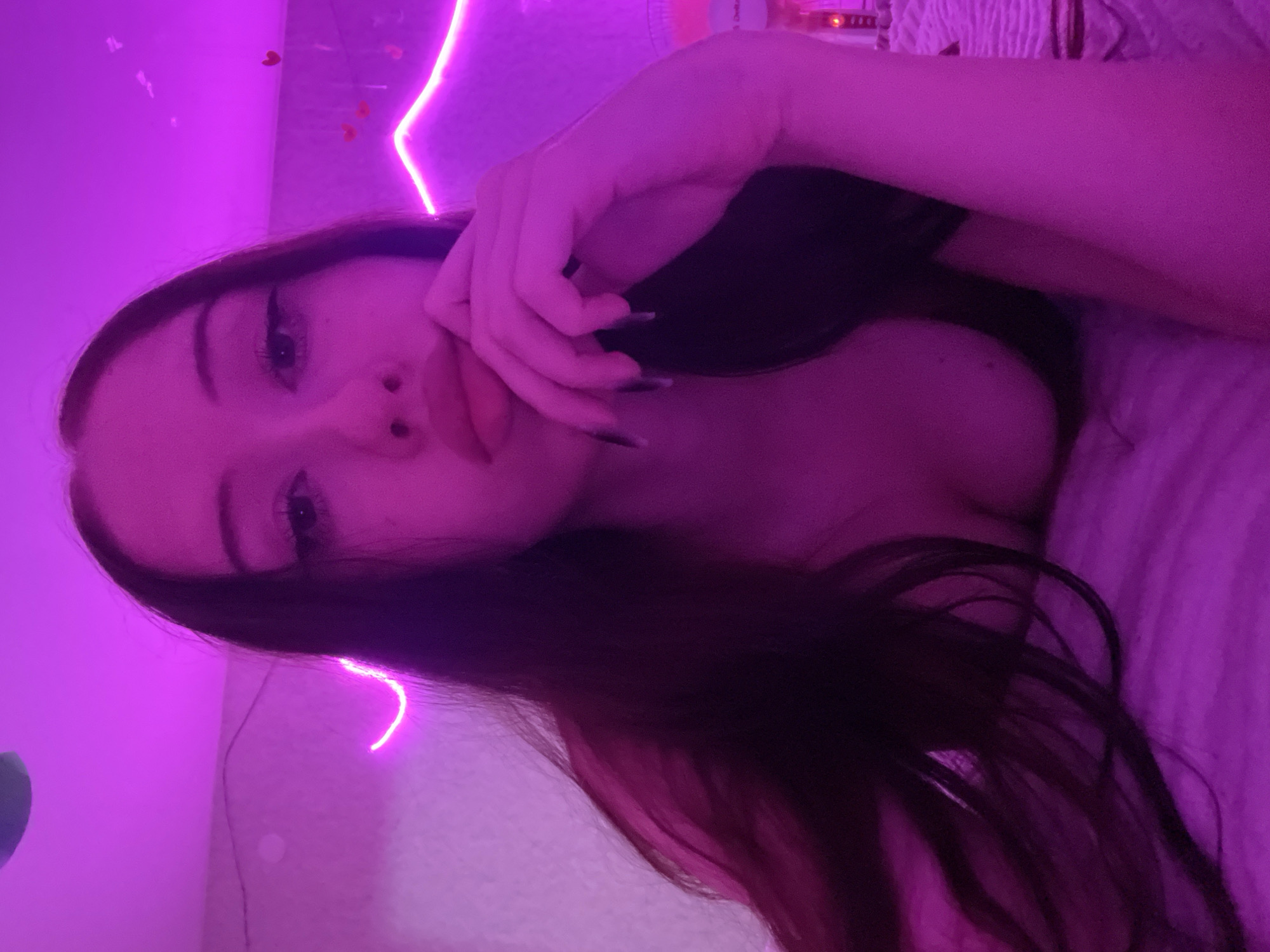 A young woman with long dark hair is lying in bed. She is illuminated by purple neon lights. She is wearing a dark top. Her left hand is resting on her face, with her fingers near her mouth. The room appears to be decorated with heart-shaped lights. The bedding is white and has a textured pattern.
