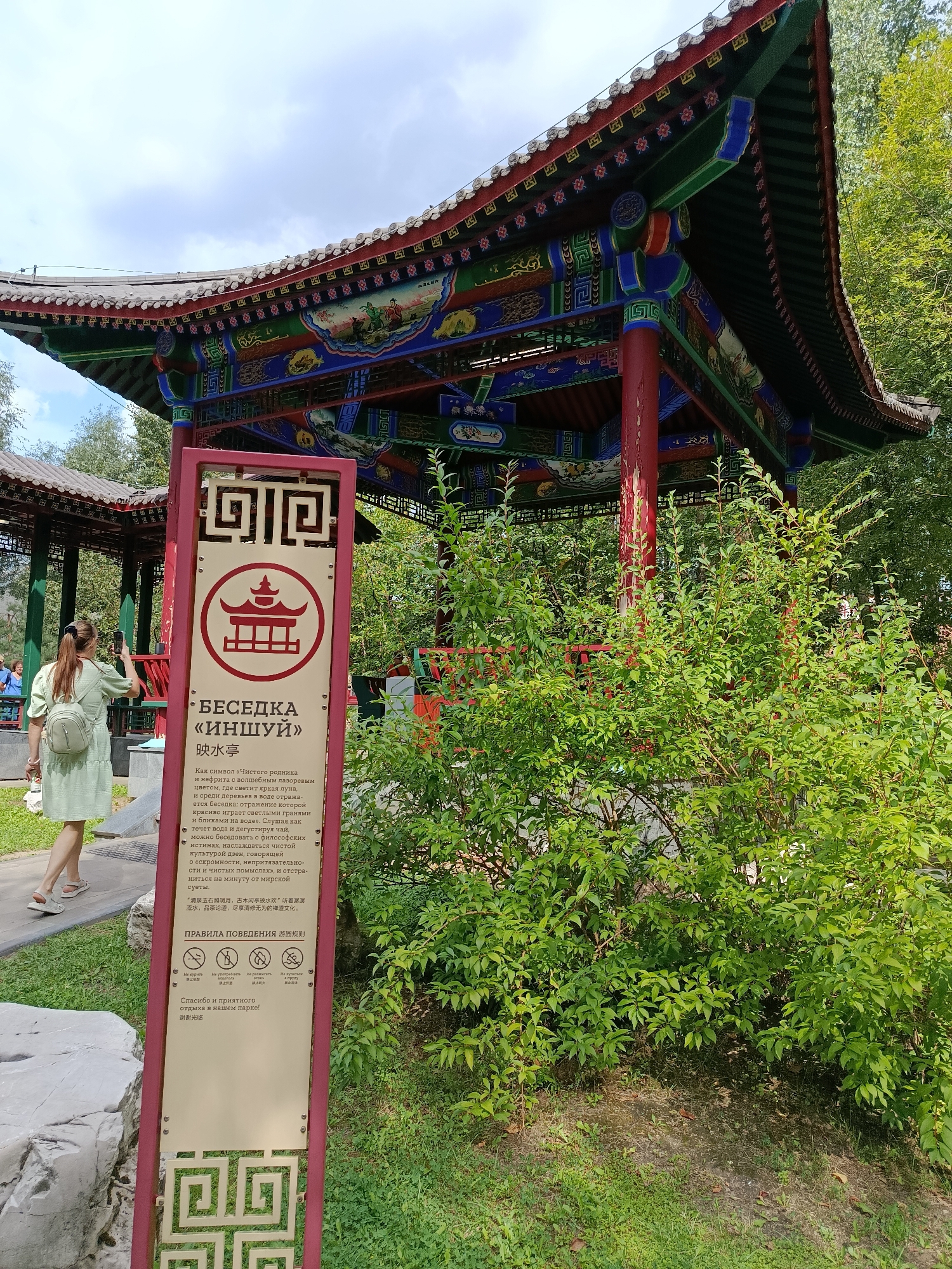 The image depicts a traditional Chinese-style pavilion in a park setting. The pavilion features a multi-tiered, brightly colored roof with intricate designs in red, green, blue, and gold. Red pillars support the roof. In the foreground, a red signpost with gold lettering and a circular emblem stands beside a lush green bush. The signpost contains text in Russian and Chinese, with icons indicating rules. A woman in a light green dress and a white backpack is visible on the left, taking a photo with her phone. The environment appears to be a park with grass, trees, and a stone. The sign indicates that this is "Besedka 'Inshui'" which translates to "Pavilion 'Inshui'" and that it is located in the Moscow City Government (MGMP) Park, also known as the Park of the Moscow Government. This location is in Moscow, Russia.