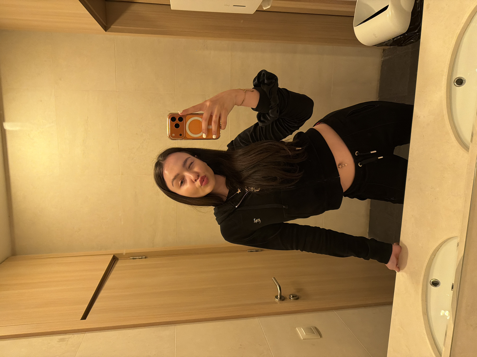 A young woman is captured in a mirror selfie within what appears to be a public restroom or bathroom. She has long dark hair and is wearing a black velour tracksuit, comprising a zip-up hoodie with a decorative gold zipper pull and matching pants. Her right arm is extended, holding an orange-cased smartphone with a prominent white MagSafe ring, angled to take the photo in the mirror. She strikes a playful pose, winking with her left eye and pouting her lips.
Notable details include a golden bracelet on her right wrist, a black scrunchie on her left wrist, and a small belly button piercing. The setting features light beige tiled walls, a light wooden cabinet or door on the left, and a light-colored countertop with a white sink visible on the right. A white hand dryer, with "DYSON" faintly visible on its surface, is mounted on the wall above the sink area, with a black trash bag underneath. The lighting is warm and artificial, suggesting an indoor environment.