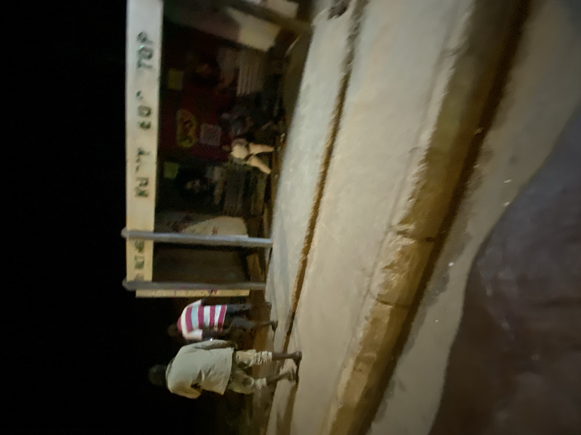 The image appears to show a street scene in Bukuru, Nigeria, at night. Two men are visible outside a building with the words "KILOTOP" on its facade. One man wears a light-colored shirt and shorts, and the other wears a striped shirt. The surrounding area is dark.