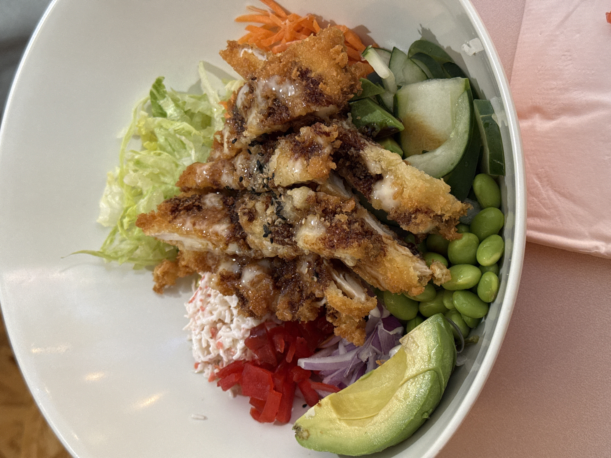 A close-up shot captures a white bowl filled with a colorful poke bowl. The bowl contains a generous portion of crispy, breaded chicken, sliced into strips and drizzled with a creamy white sauce. Surrounding the chicken are various fresh ingredients, including shredded lettuce, shredded imitation crab meat with a pink hue, bright red pickled ginger, thinly sliced purple onion, creamy avocado slices, edamame, shredded carrots, and sliced cucumber. The poke bowl is presented on a light pink textured surface, likely a tablecloth or napkin. The lighting in the scene is bright, highlighting the textures and vibrant colors of the food. The image appears to be taken indoors, possibly in a restaurant setting, though no specific location within Tacoma, United States of America can be identified from the visual information provided.