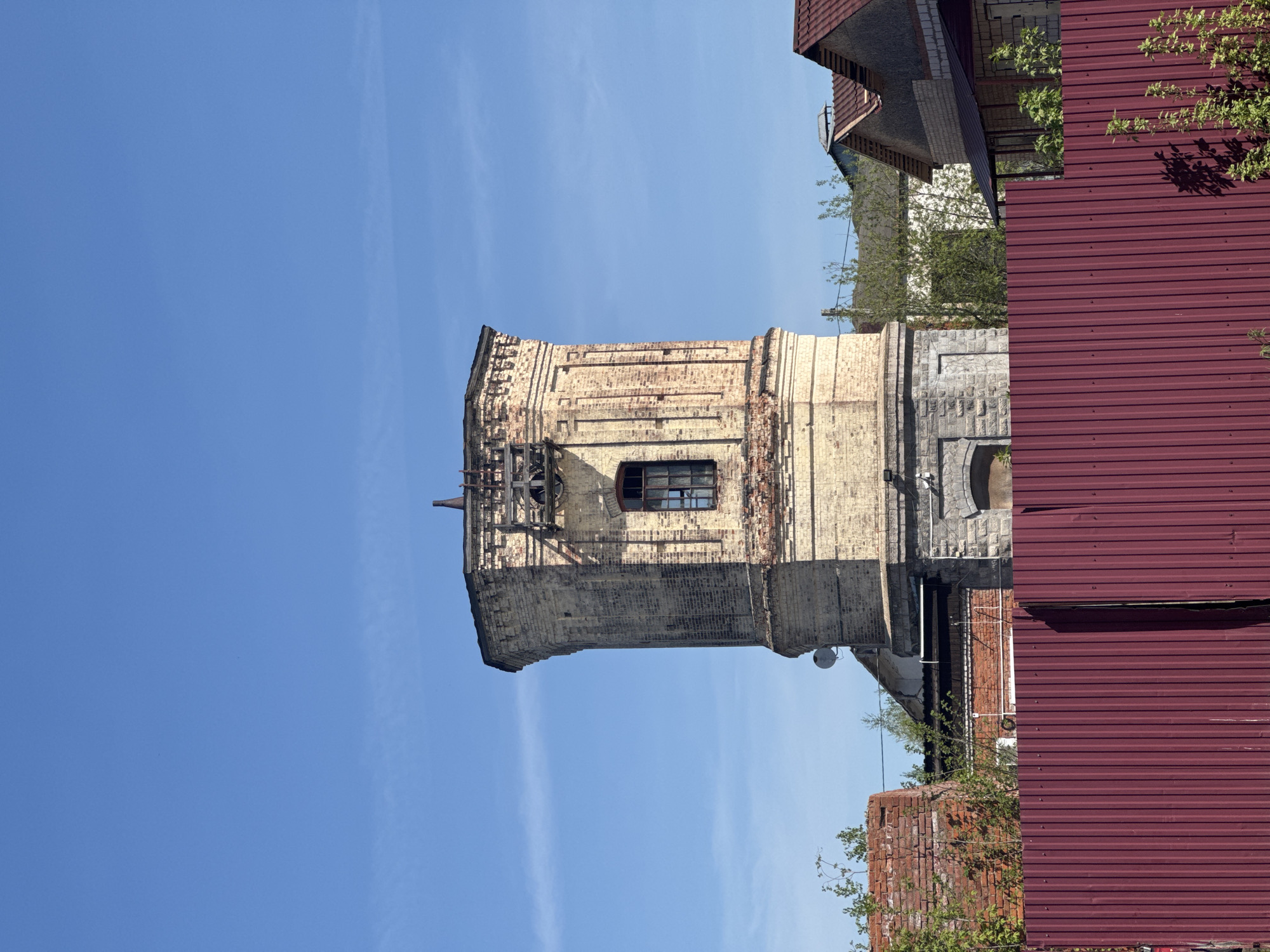 The image depicts an old, partially dilapidated brick tower under a clear blue sky. It's likely the former water tower in Balezino, Russia, judging by its architectural style and the presence of similar structures in the region. The tower is mostly constructed of yellow brick with visible signs of weathering and damage, including missing bricks, particularly near the top. There's a dark window visible on one side of the tower. Wooden supports and scaffolding are present on the upper part of the tower, suggesting ongoing repairs or structural deterioration. Parts of nearby buildings are visible, including a section of a roof covered with red corrugated metal sheets and a building with a combination of brick and stone walls. Some greenery, potentially trees or bushes, is present in the foreground and around the tower.