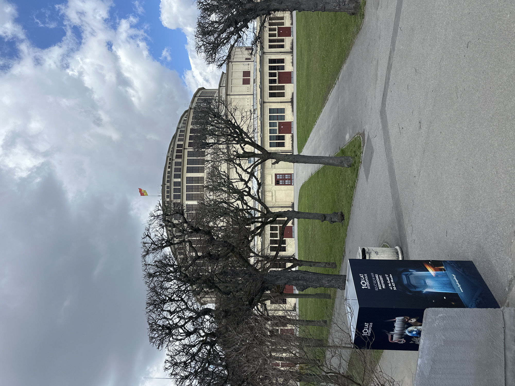 The image shows a side view of the Centennial Hall in Wrocław, Poland, partly obscured by bare trees. A pathway and grassy areas extend from the building. In the foreground is a large black rectangular advertisement for Hydropolis, an interactive science museum in Wrocław, located at ul. Na Grobli 17. The sky is overcast, with patches of blue visible.