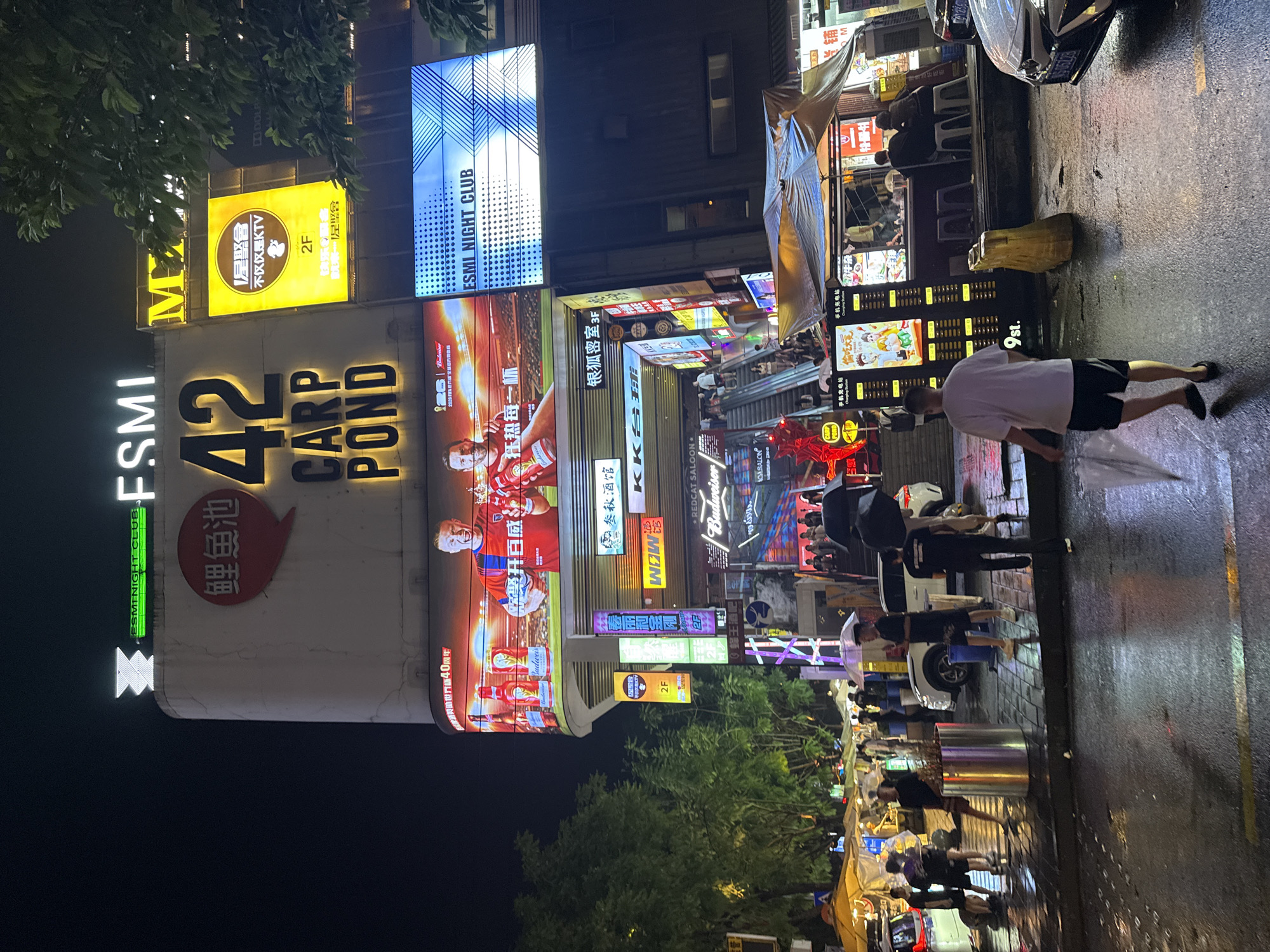 The image depicts a nighttime street scene in Liangjiang, China, characterized by bright neon signs and wet pavement, suggesting recent rain. A large building with multiple illuminated advertisements dominates the background. Prominent signage includes "42 CARP POND" in large yellow letters, a Budweiser advertisement featuring soccer players, and "ESMI NIGHT CLUB" displayed on a blue and white LED screen. Smaller signs indicate various establishments like "Mizunoi Wine House" and "KKK Stadium."
In the foreground, several individuals are walking on the wet sidewalk. Some are holding umbrellas, further emphasizing the rainy conditions. One person in a white t-shirt and black shorts is walking away from the camera, holding a translucent umbrella. Several white and black cars are parked along the street. The street level also features food stalls and what appears to be a charging station. The overall atmosphere is bustling and vibrant, typical of an urban nightlife area.