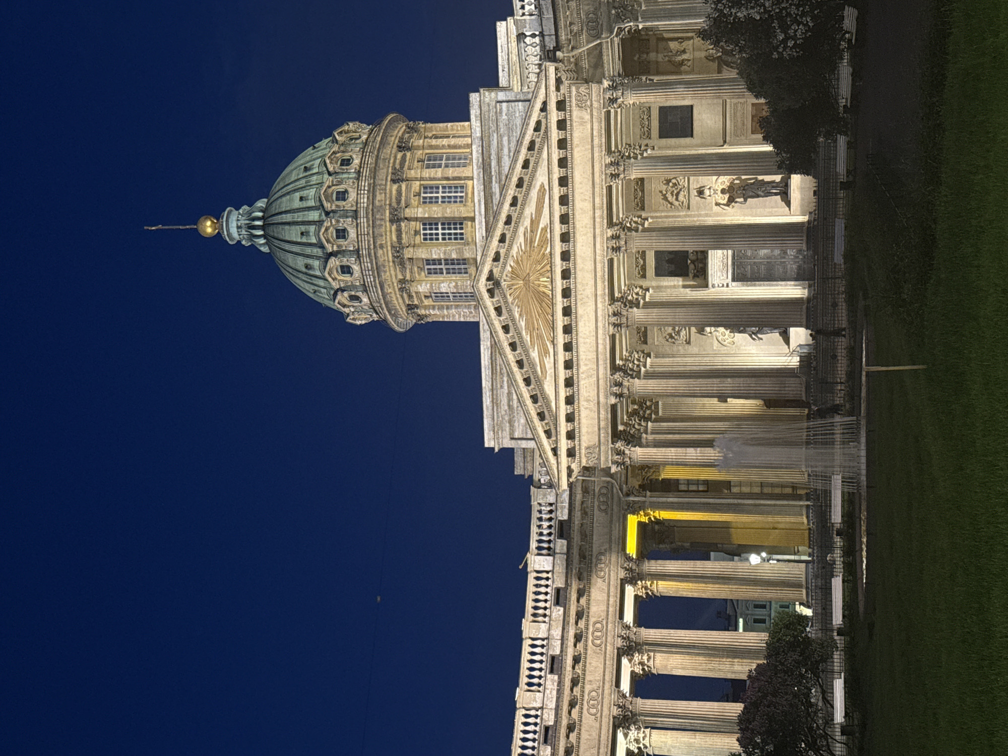 The image captures the Kazan Cathedral in Saint Petersburg, Russia, at night or deep twilight, set against a clear, dark blue sky. The grand, classical edifice dominates the frame, illuminated by warm artificial light that highlights its intricate details.
The cathedral features a distinctive, large green dome, topped with a golden finial and a cross. Below the dome, a circular drum with multiple arched windows is visible. The main facade is characterized by an extensive colonnade of tall, classical columns, likely Corinthian, supporting a detailed entablature. A prominent pediment surmounts the central section, adorned with a radiant relief sculpture, appearing as a sunburst or an Eye of Providence motif. Numerous statues are positioned along the facade, standing between columns and in niches, adding to the building's classical grandeur.
The building's stone exterior glows with yellow and white light, accentuating its architectural features and textures. Some windows within the dome and facade emit a soft, warm light from within.
To the right of the cathedral (in a rotated view, this would be the ground level), a well-maintained grass lawn is visible, interspersed with trees and bushes. Several white park benches are placed along a pathway, indicating a public space or square adjacent to the cathedral. A fence separates the green area from the building. One cropped image also reveals what appears to be a fountain with water jets. No specific activity is taking place, and no people or legible text are clearly visible. The scene conveys a sense of serene majesty and architectural beauty under the night sky.