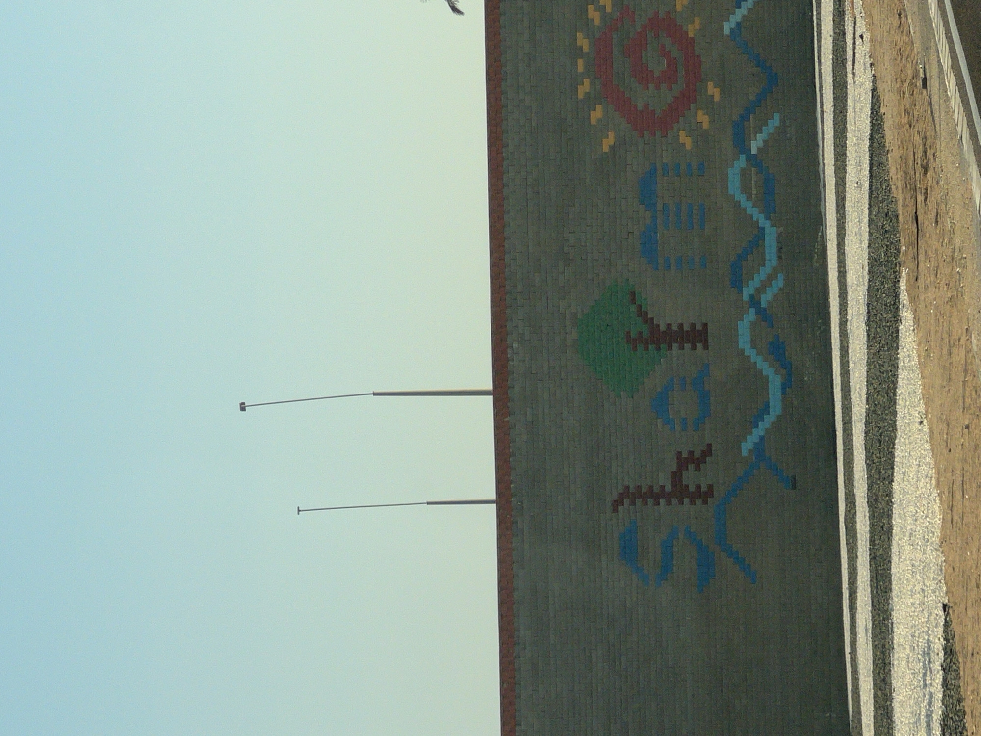 The image shows a brick wall decorated with a mosaic. The mosaic depicts the word "Sharm" in stylized blue and red letters, accompanied by a green tree on the left and a red and yellow sun on the right. Below the word, there is a wavy blue pattern. The wall is topped with a band of red bricks. In the background, two tall light poles are visible against a clear blue sky. The foreground shows a path made of white and dark grey gravel, with a sandy area to the right. The overall scene suggests a public space or a decorative element within the city of Sharm El Sheikh, Egypt.