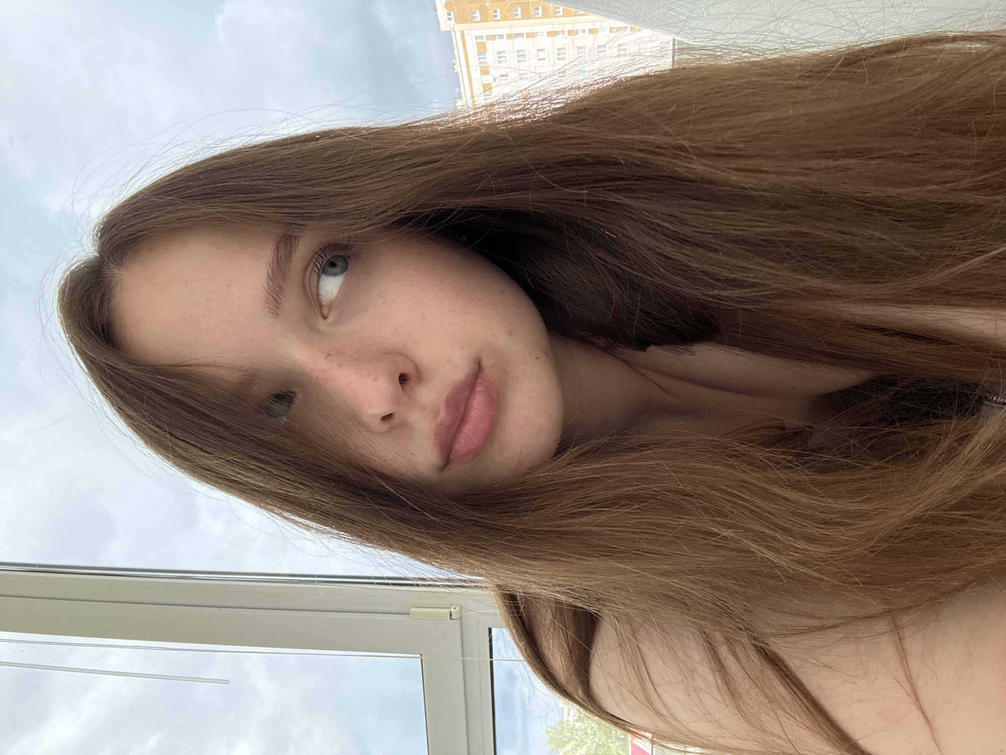 A young woman with long brown hair and freckles is pictured in a selfie. She is looking to her right, with her mouth slightly parted. The background includes a window with vertical blinds on the left and a building with multiple windows on the right, suggesting an urban setting. The sky appears cloudy. The image is taken from a medium close-up, focusing on her face and upper body. The setting is likely an apartment or room in Chaikovsky, Russia, given the building architecture visible.