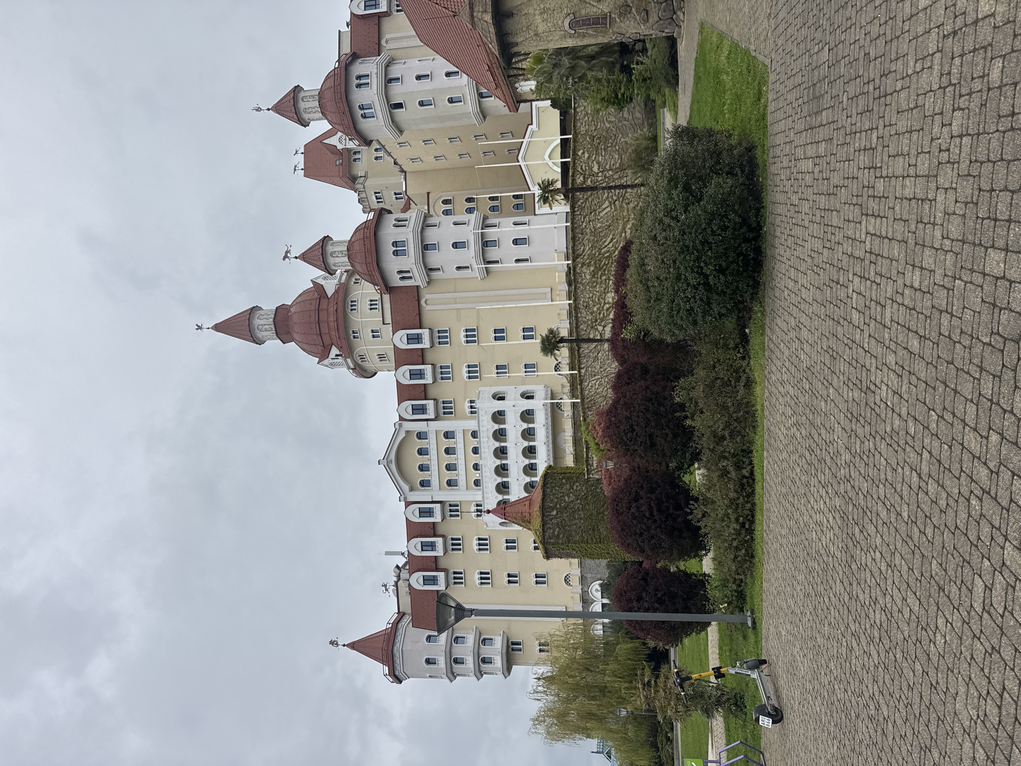 An outdoor, slightly rotated image captures a large, castle-like building under an overcast sky, situated in a landscaped setting with a paved walkway. The multi-story building is predominantly light beige with numerous distinct turrets topped with red-tiled conical roofs. Many windows of various shapes, including arched and rectangular, are visible across its facades. Several flagpoles stand in front of the building, and some sections of its walls are covered in dense ivy or climbing plants. Distinctive weather vanes, possibly depicting animal or mythical figures, crown several turrets.
In the foreground, a wide pedestrian path made of grey cobblestones or pavers stretches across the scene. Adjacent to the path and in front of the building are well-maintained green spaces, featuring manicured lawns, dense bushes with both dark green and reddish-purple foliage, and several trees, including palm trees. An electric scooter, colored yellow and grey, is parked on the edge of the paved path in the lower right corner, bearing the partial text "AREN" and a number plate "490 90". Street lamps are also visible within the scene.
The weather appears to be a uniformly grey, cloudy day with diffused lighting. No people are present, lending a quiet atmosphere to the scene. The architecture strongly suggests a resort or themed hotel, consistent with its location in Sirius, Russia, likely depicting the Sochi Park Hotel.