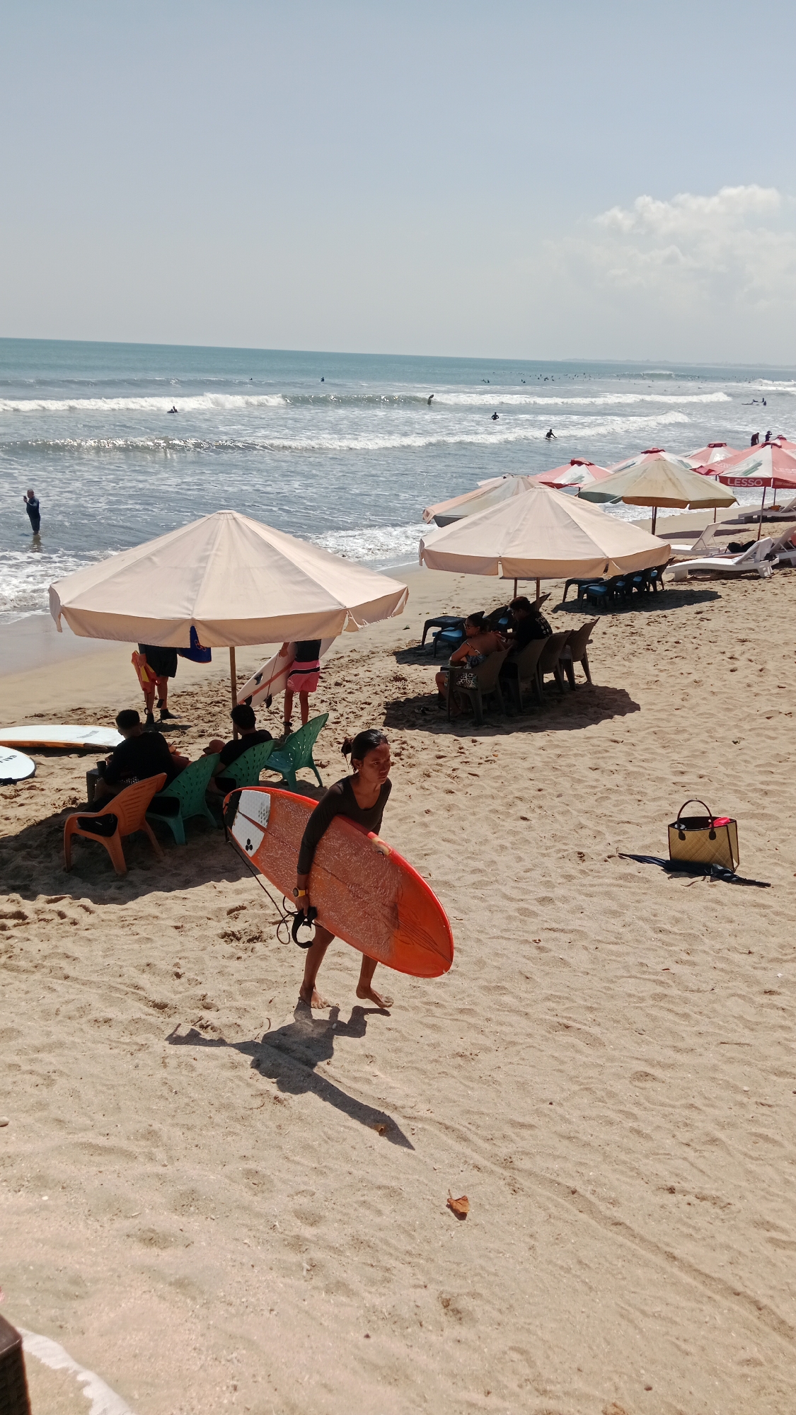 A medium-shot image captures a sunny day at a beach with many people and surfboards. The foreground features a woman walking on the sand, holding a bright orange surfboard with a white pattern. She is wearing a dark long-sleeved shirt and dark bottoms. Her shadow stretches across the sand. Behind her, several beach umbrellas are set up, providing shade for groups of people sitting on chairs and loungers. Some individuals are visible in the water, with surfboards, suggesting they are surfing. The ocean is calm with gentle waves, and the sky is clear with a few clouds. A straw bag with red handles rests on the sand to the right of the frame. The overall scene depicts a lively beach environment with recreational activities.