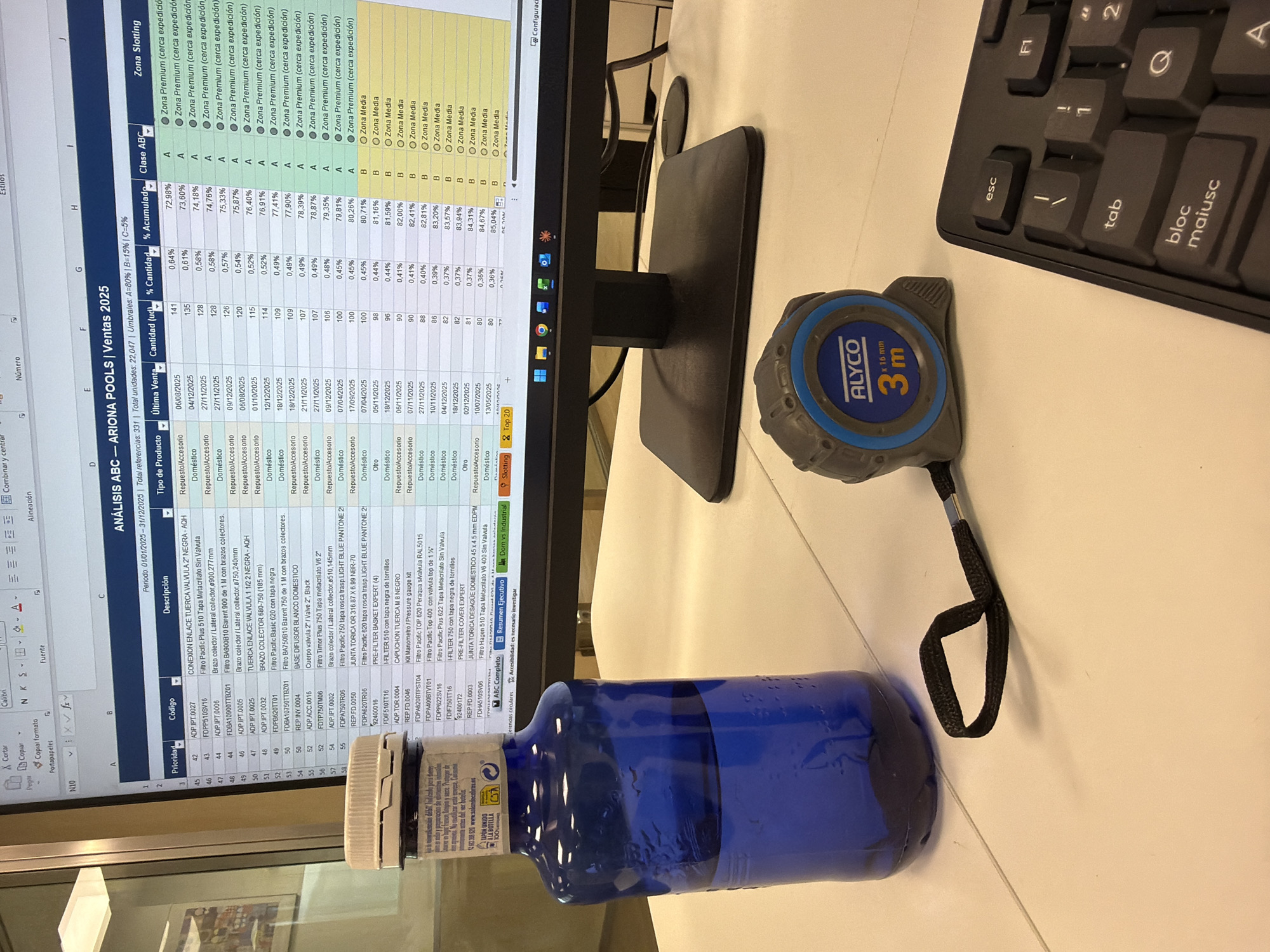 The image shows a desk with various items on it. In the foreground, there's a blue bottle, a blue and grey measuring tape marked "ALYCO 3m", and a black keyboard. Behind these items is a computer monitor displaying an Excel spreadsheet. The spreadsheet appears to be an analysis of "ARIONA POOLS | Ventas 2025" with details on product codes, descriptions, sales data, and zone classifications. The location in the city of Errenteria, Spain, is not discernible from the visual information provided.