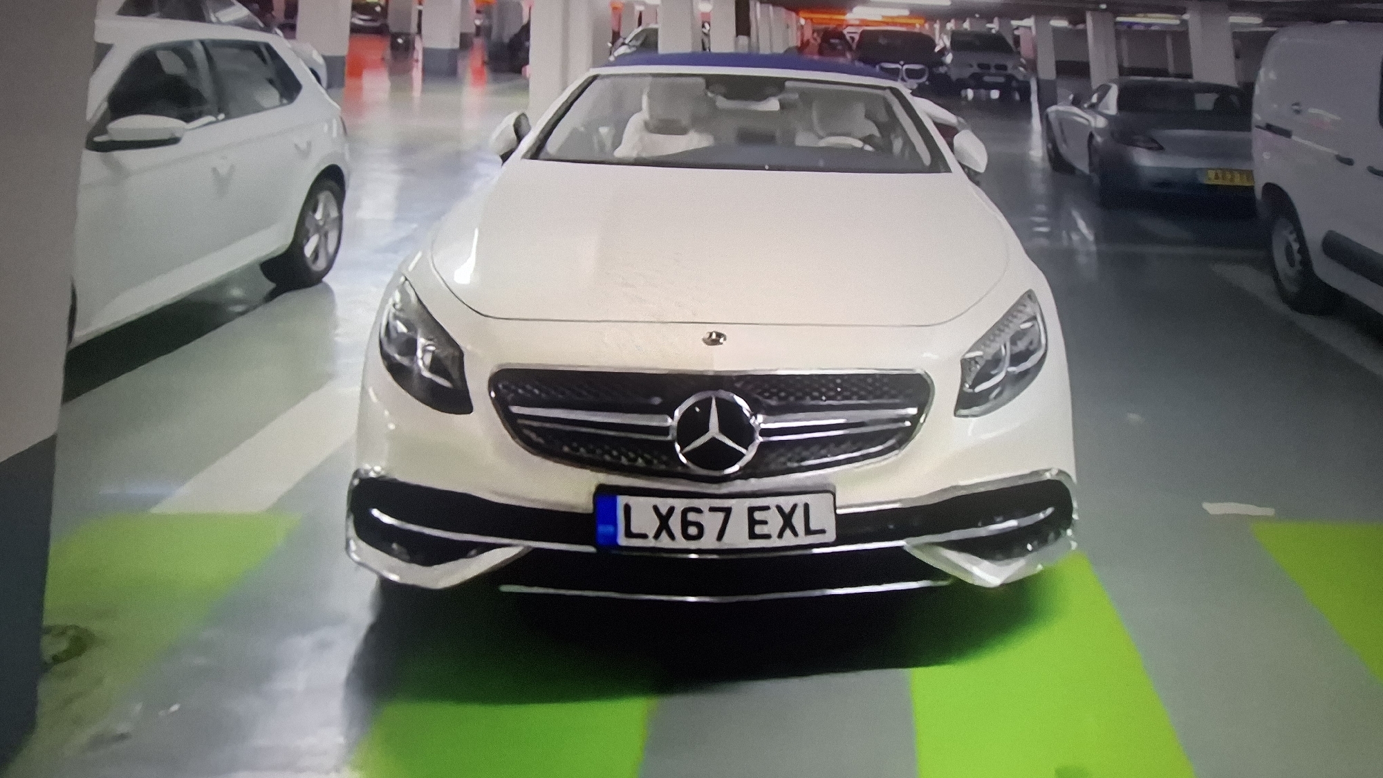 The image depicts a white Mercedes-Benz S-Class convertible with the license plate "LX67 EXL" parked in an indoor parking garage. The car is facing the camera, centered in the frame. Other cars are visible in the background, including a white hatchback on the left, a grey sports car and a white van on the right. The garage has concrete pillars and a grey floor with a green parking space marker under the Mercedes. The location is not identifiable.