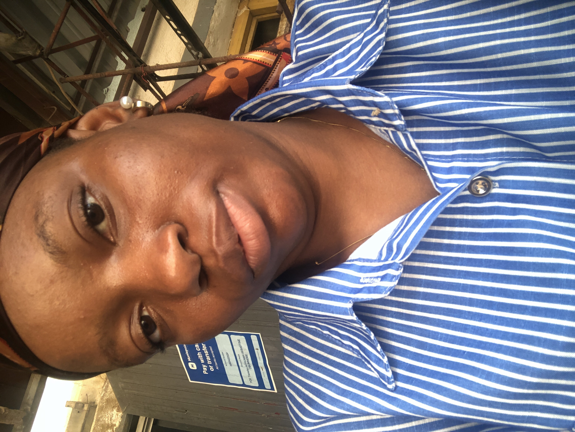 A medium shot shows a woman from the chest up, wearing a blue and white striped button-down shirt. A brown patterned headscarf covers her hair. A pearl earring is visible. The woman has a dark complexion. She is looking directly into the camera with a slight smile. Behind her is a partially visible metal structure. A sign reading "Moniepoint, Pay with cash or transfer" is visible in the lower left corner. This suggests the location may be a business or point of sale. The sign includes the text "POS Transfer-ACHUAMA SEBASTINE OBUMNEME," implying that the location is in Nigeria, although the exact city, Jos, cannot be determined from the image.