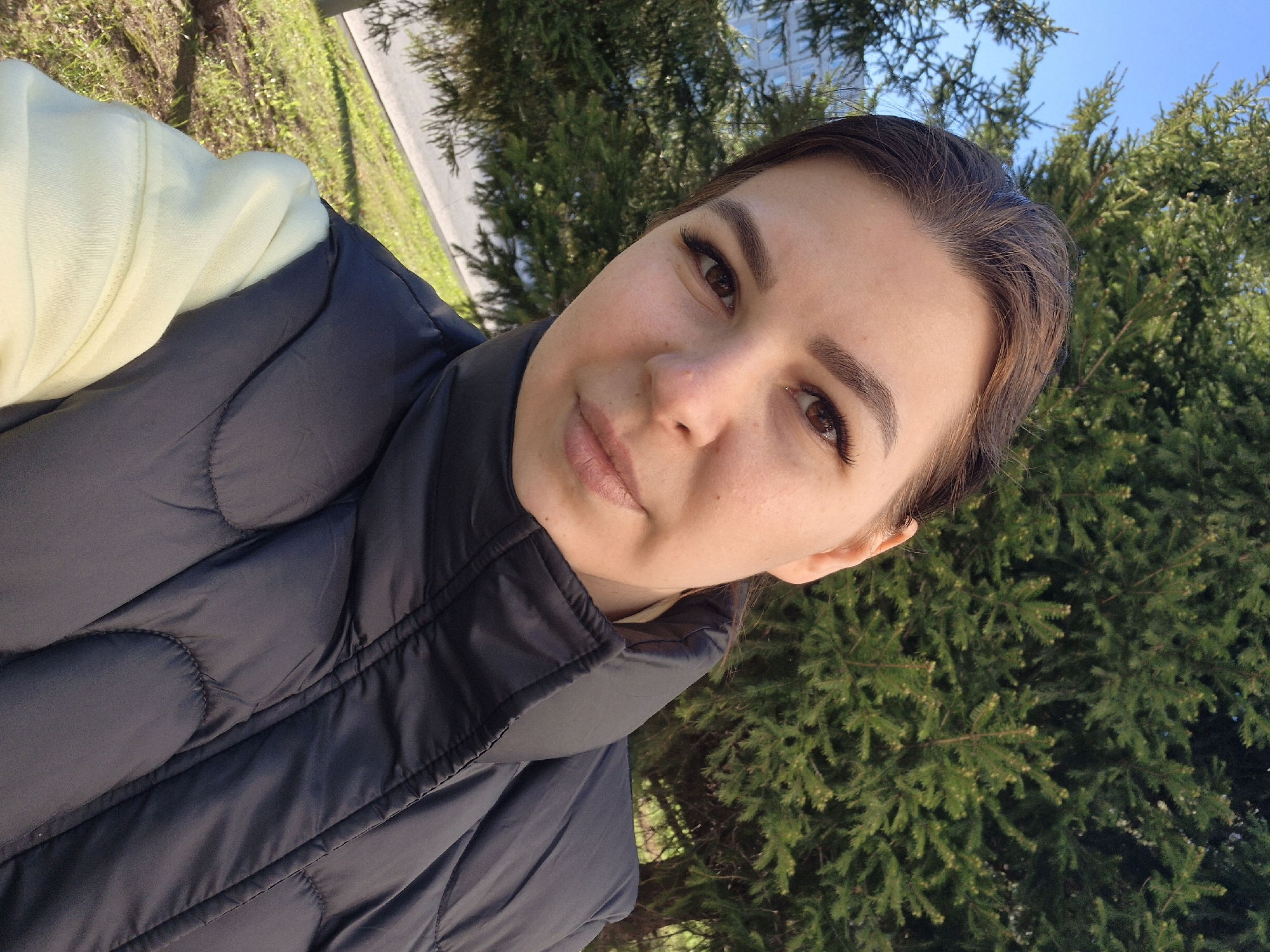 A person with brown hair, brown eyes, and light skin is looking at the camera. The person is wearing a black puffy vest over a light yellow garment. The background includes a green evergreen tree and a blue sky. The location is Tobolsk, Russia.