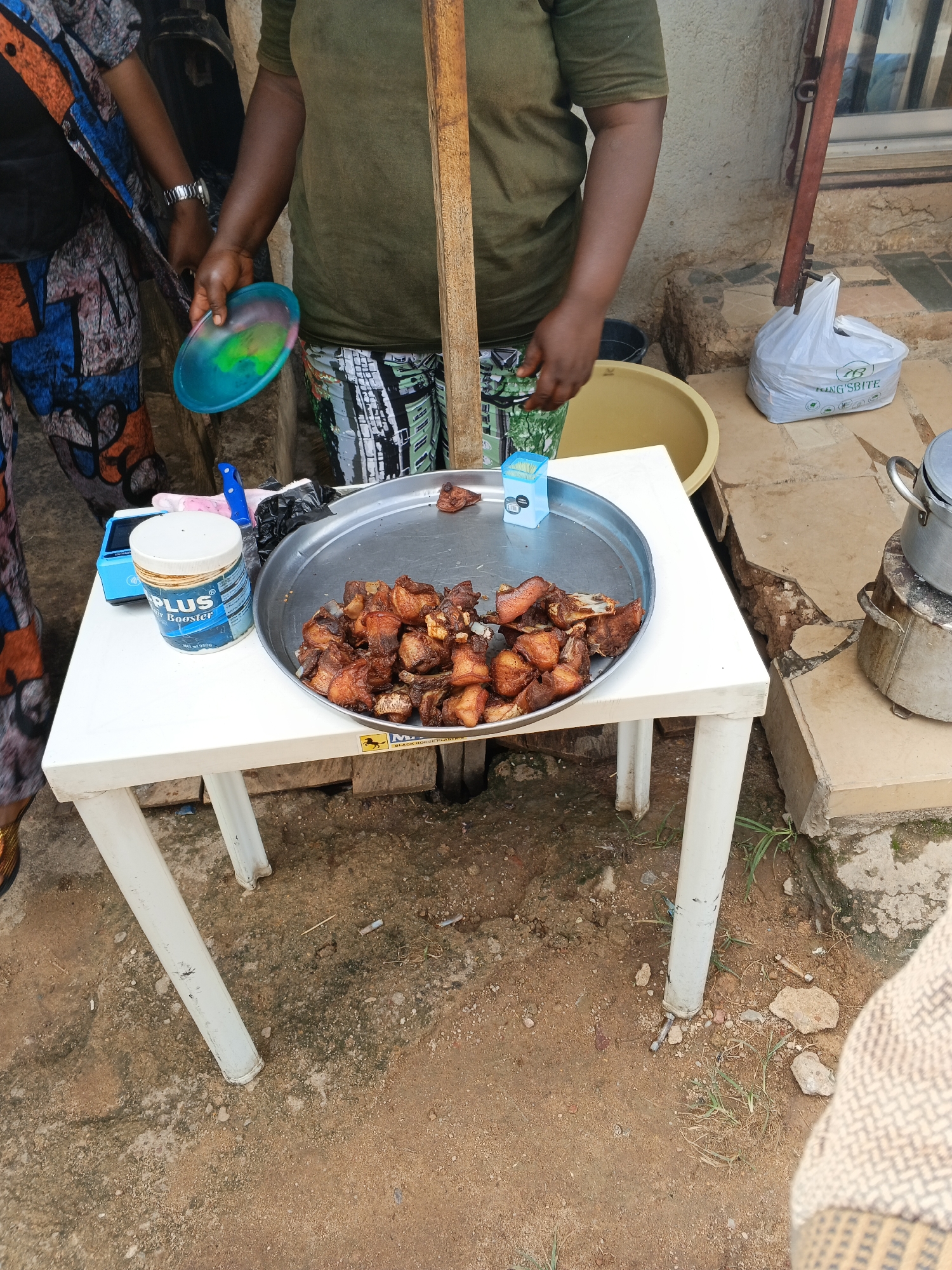 The image depicts an outdoor scene, likely in Jos, Nigeria, where a person is standing behind a white plastic table. The person, whose lower body is visible, is wearing a green t-shirt and patterned trousers. They are holding a colorful plastic plate in their left hand and reaching towards a metal tray filled with fried meat.
On the table, in front of the tray of food, is a blue and white container labeled "PLUS Air Booster" with text indicating "Net Wt. 250g". Next to it, a small blue box of toothpicks is also visible. A large yellow bowl is partially visible behind the table.
The background shows a textured wall, a wooden pole, and some debris on the ground. To the right of the table, a white plastic bag with text that includes "KING'SBITE" is placed on the ground, next to a metal cooking pot on a stand. The ground is a mix of dirt and small stones, with some patches of dry grass. The lighting suggests it might be daytime, with overcast or diffused light. The overall scene suggests a casual outdoor food preparation or serving situation.