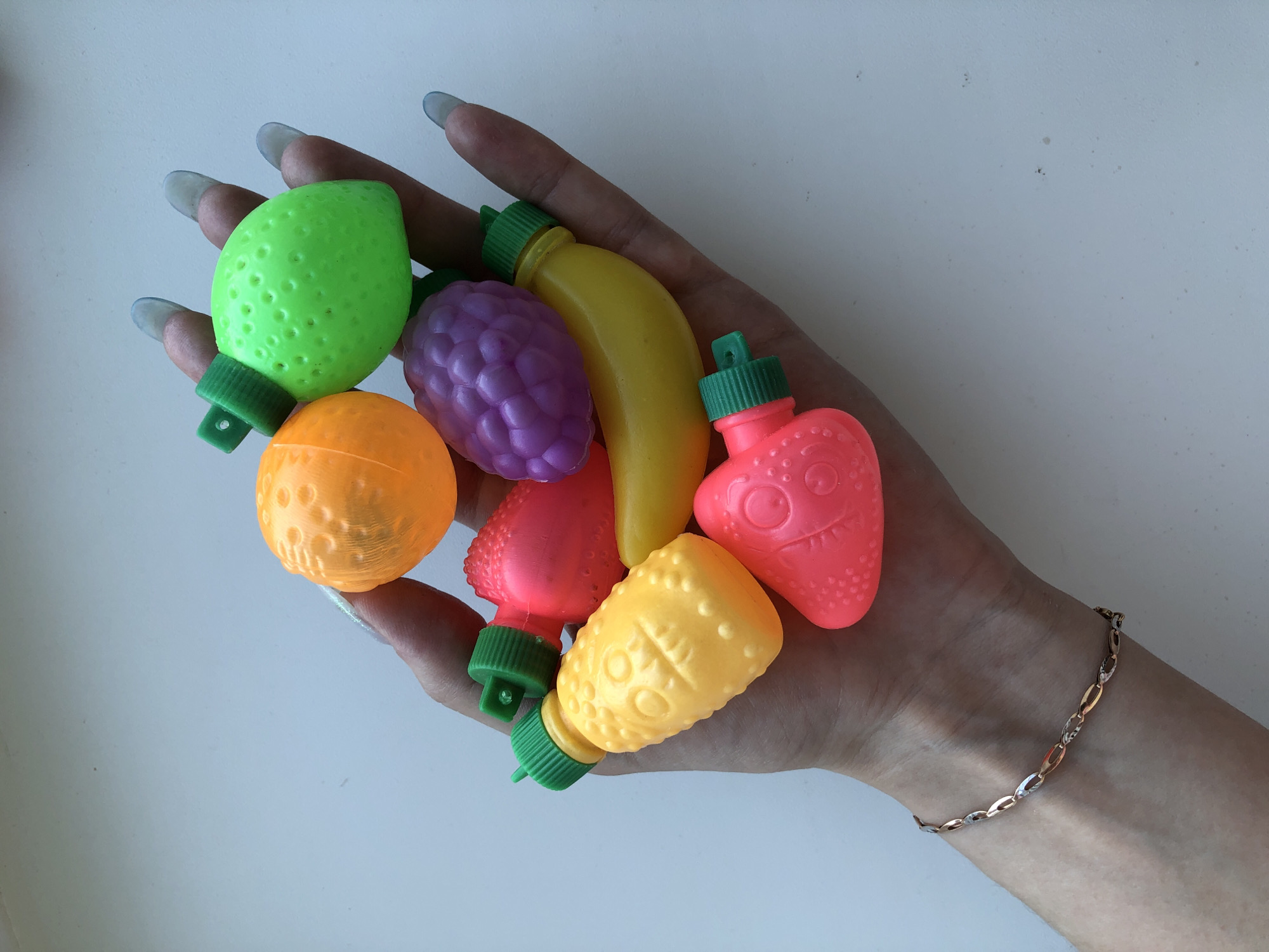A fair-skinned human hand, adorned with long, translucent acrylic nails, cradles a collection of plastic toy fruits. The assortment includes a green lemon, orange, purple grapes, yellow banana, red strawberry, and yellow corn cob. Each fruit is affixed with a green plastic cap. A gold bracelet encircles the wrist. The backdrop is a plain white surface. The location is not discernible from the image and cannot be determined to be in Noviy, Russia.